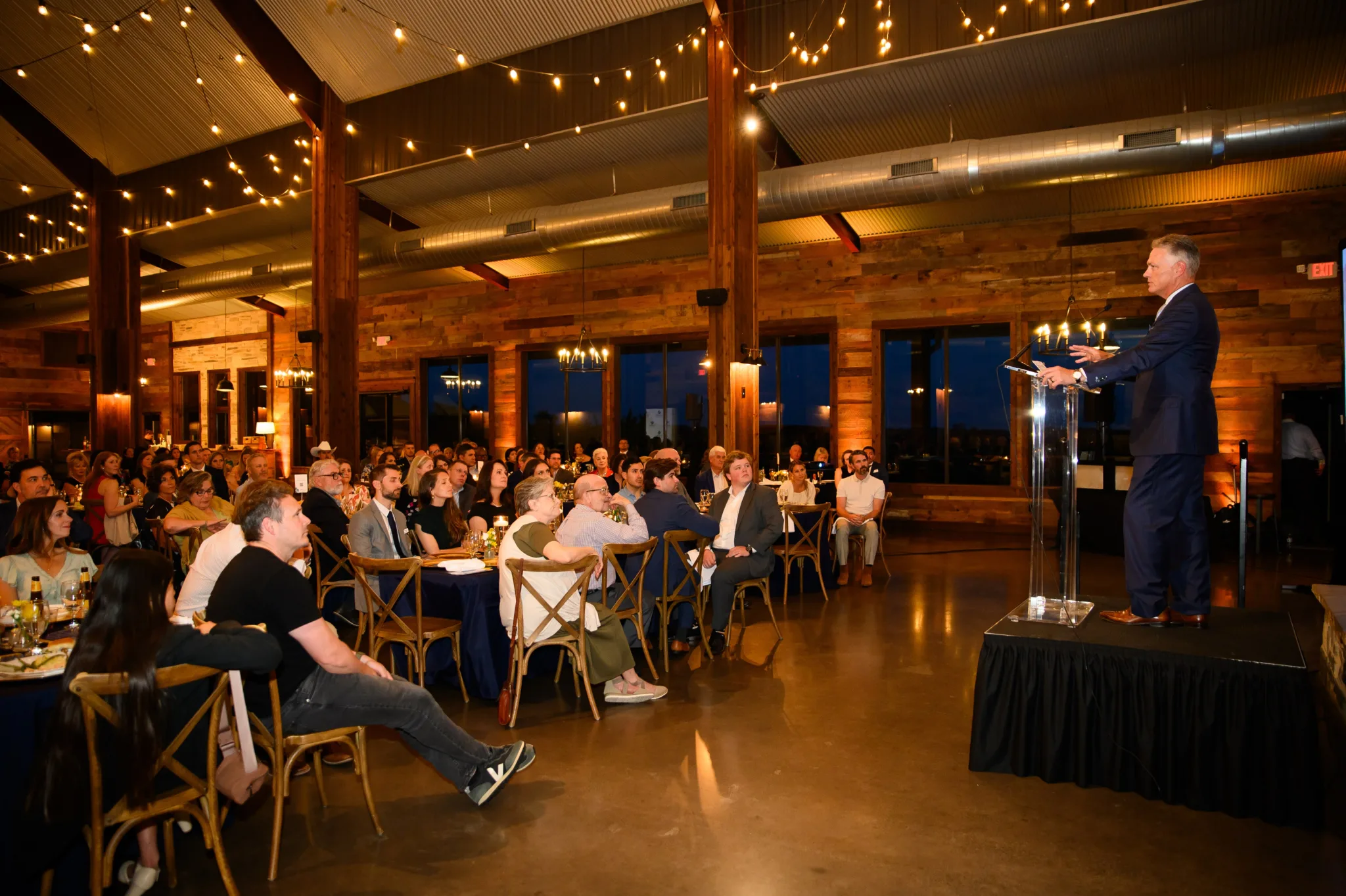 A man in a dark suit is speaking at a podium during an indoor event with a large audience seated at tables. The room has wooden walls, string lights, and large windows, with a warm lighting atmosphere.