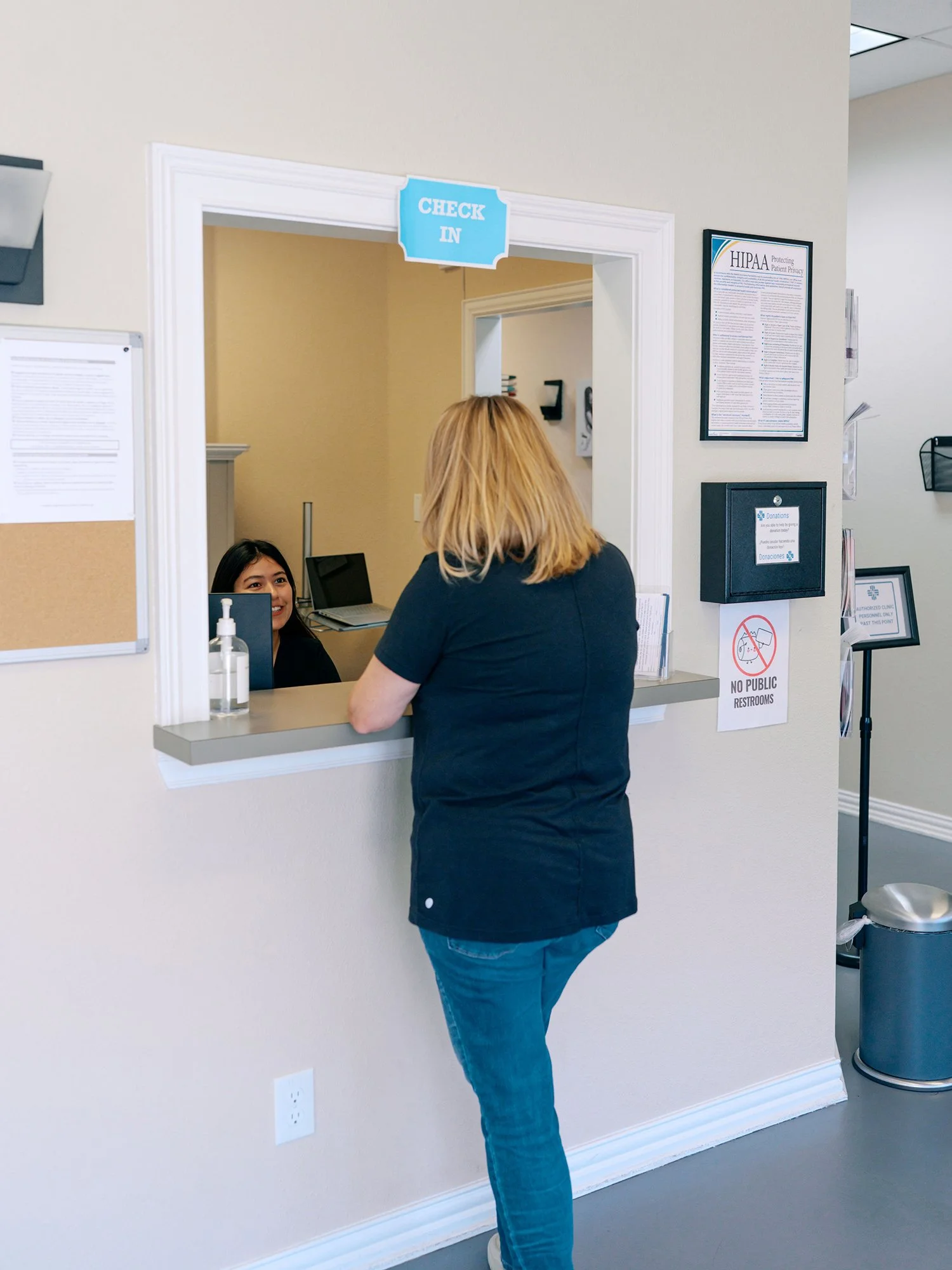 A woman with blonde hair wearing a navy blue top and jeans is at the reception window, talking to a receptionist behind the counter in a healthcare or clinic setting.