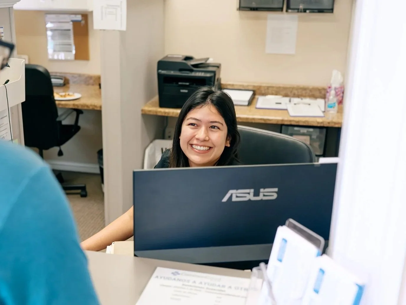 A woman smiling at a man across a desk in an office setting, with a computer, paperwork, and office equipment visible.