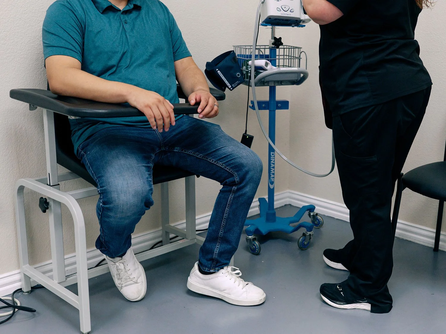 A man in a teal shirt and jeans sitting in a medical examination chair, with a healthcare worker in black scrubs nearby, adjusting medical equipment on a stand.
