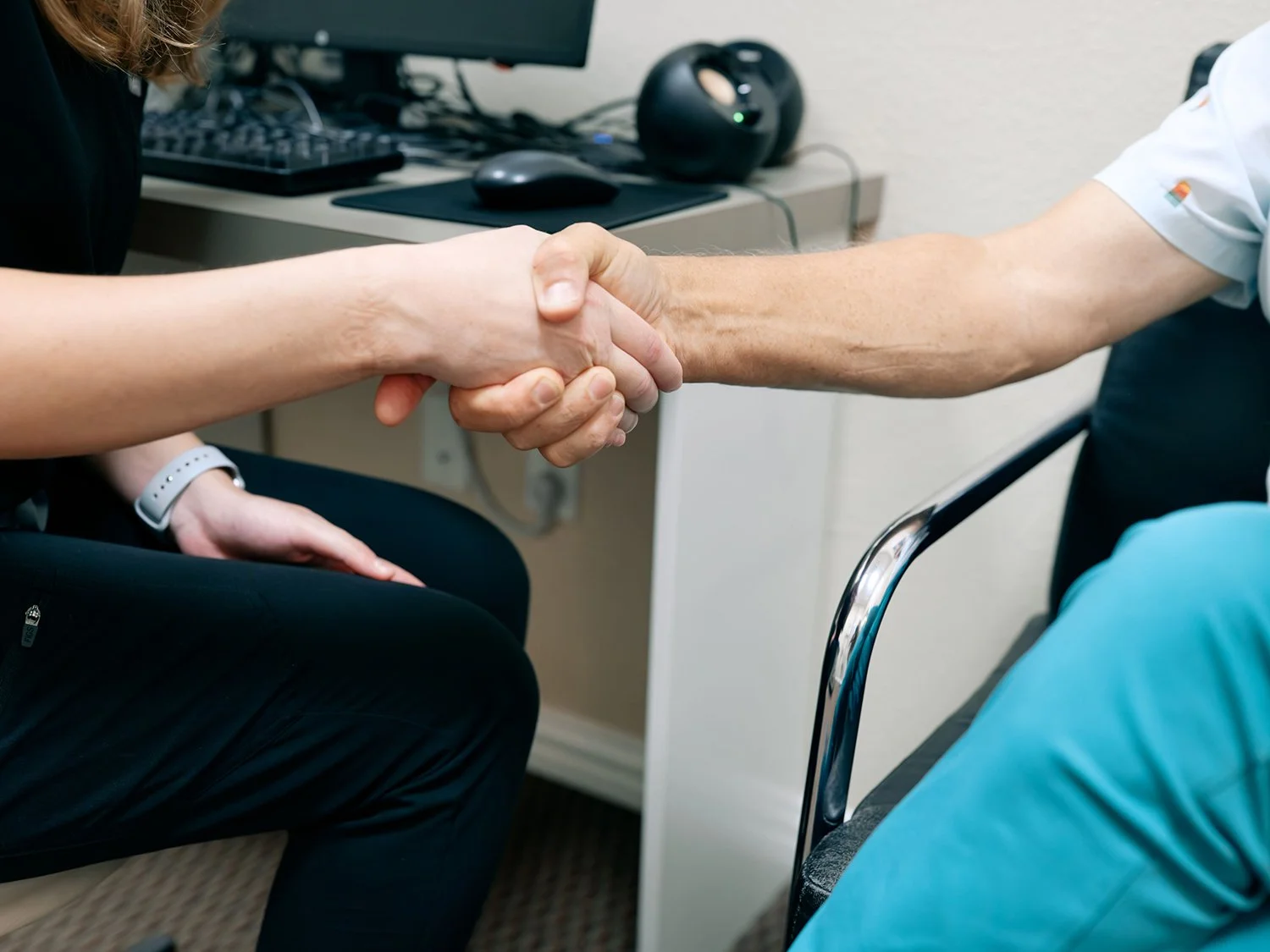 Two people shaking hands, one appearing to be a healthcare professional and the other a patient, in a medical office setting.
