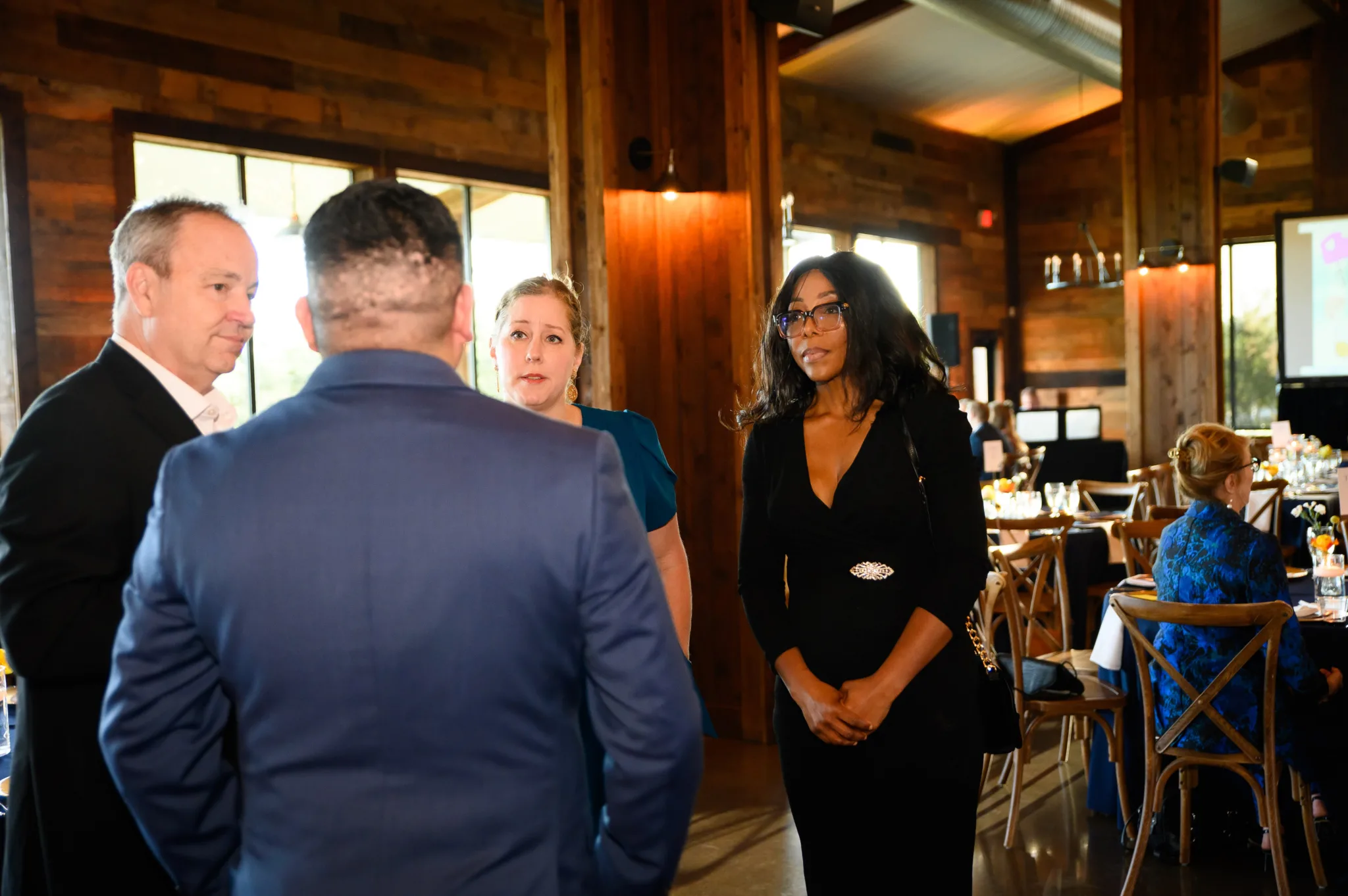 A group of four people engaged in conversation in a well-lit, wooden interior space, possibly a restaurant or event venue. There are tables with decorations and a poster or screen in the background.