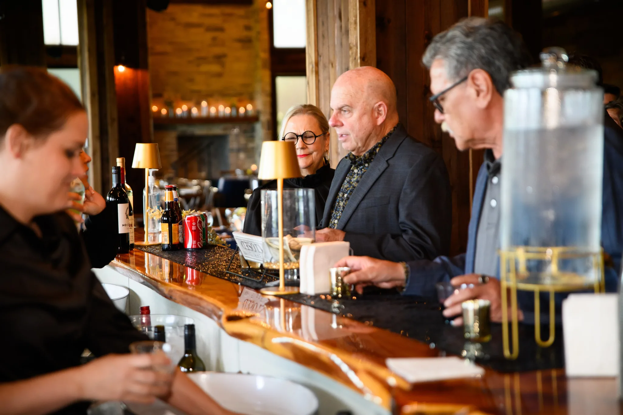 People standing at a bar counter with drinks, including wine bottles and soda cans, in a cozy, warmly lit restaurant with wooden walls and soft lighting.