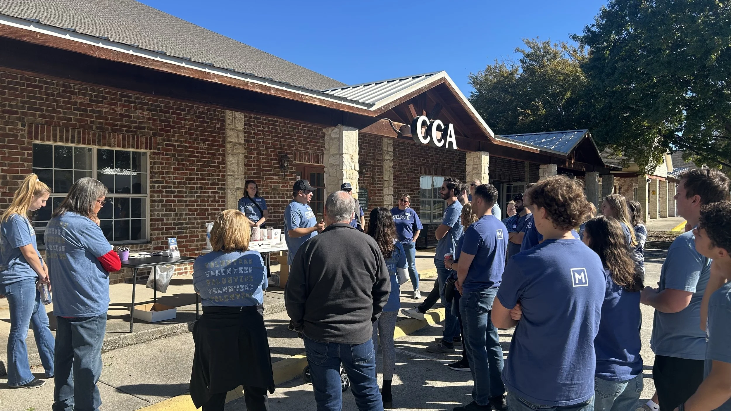 A group of volunteers gathered outside a brick building with the sign 'CCA' on the front, participating in an organized community event on a sunny day.