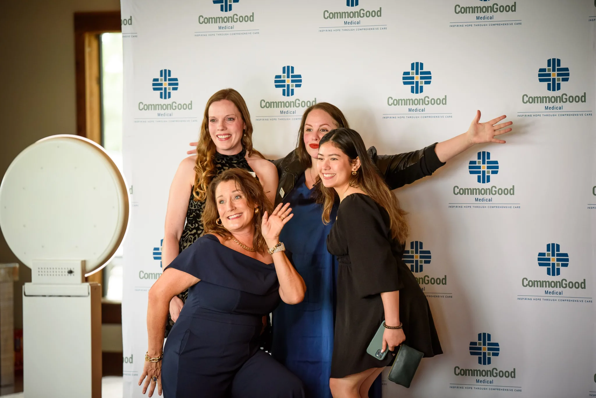 Four women posing together at a photo event in front of a backdrop with the 'CommonGood Medical' logo, with one woman making a playful gesture and another holding a smartphone.