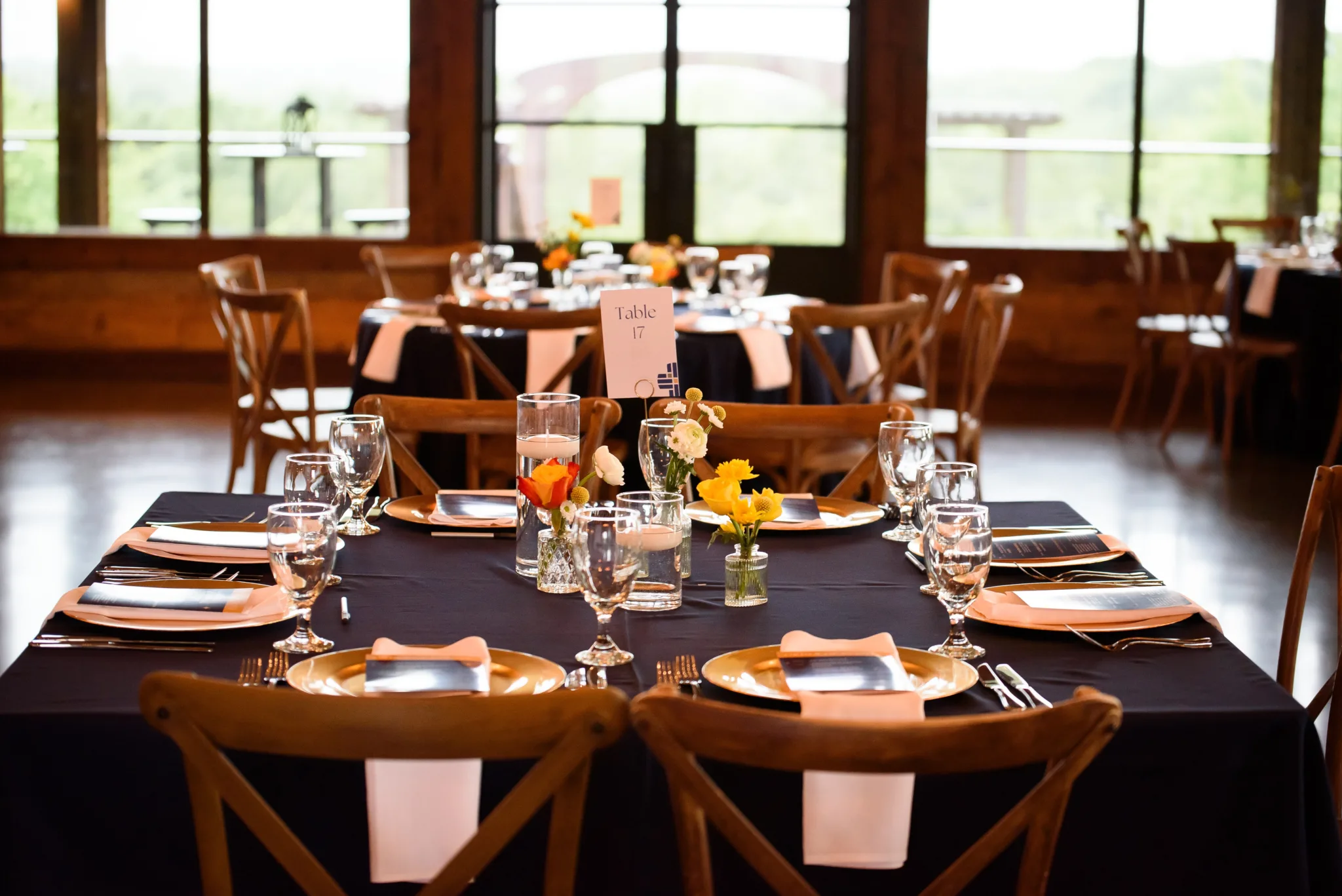 A dining table set for a celebration with a black tablecloth, gold charger plates, pink napkins, and clear glasses. The centerpieces include small vases with white, yellow, and orange flowers. A table number sign reads 'Table 17'. In the background, 