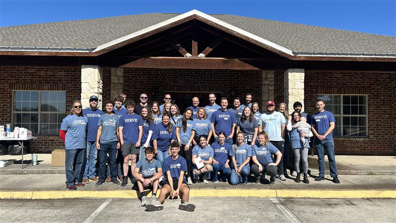 Group of people, including men, women, and children, wearing blue shirts with the word 'SERVE' standing outside a brick building.