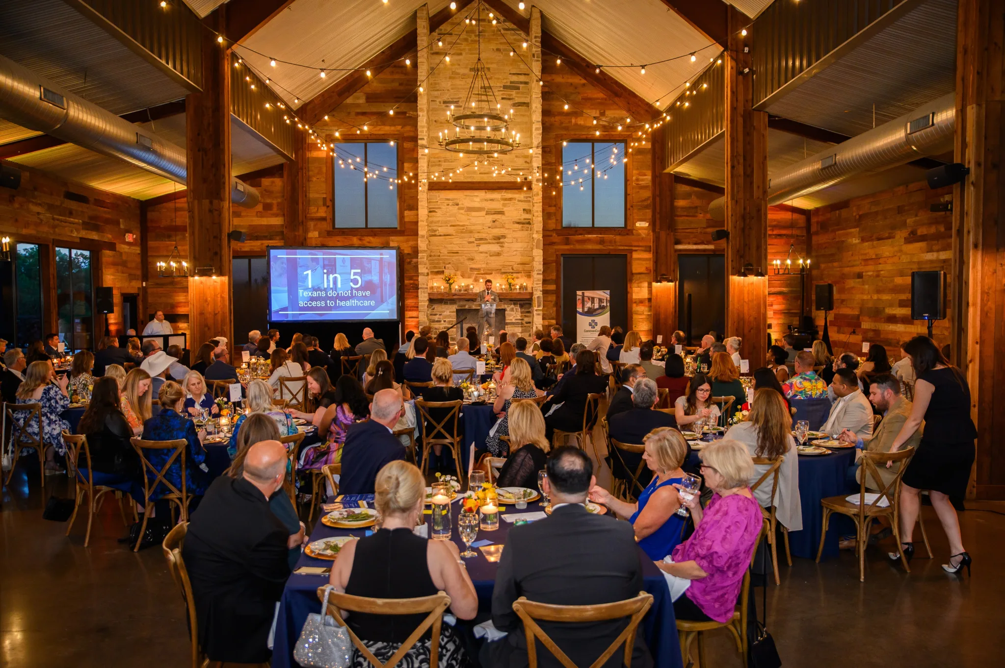 A large indoor event in a rustic-style venue with wooden walls and ceiling, featuring string lights hanging from the ceiling. There are numerous round tables with people seated, eating and talking. A large screen displays a statistic about Texans and
