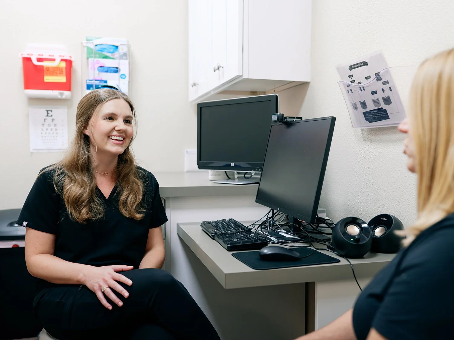 Two women in medical scrubs smiling and talking in a clinical office setting with computers and medical equipment.