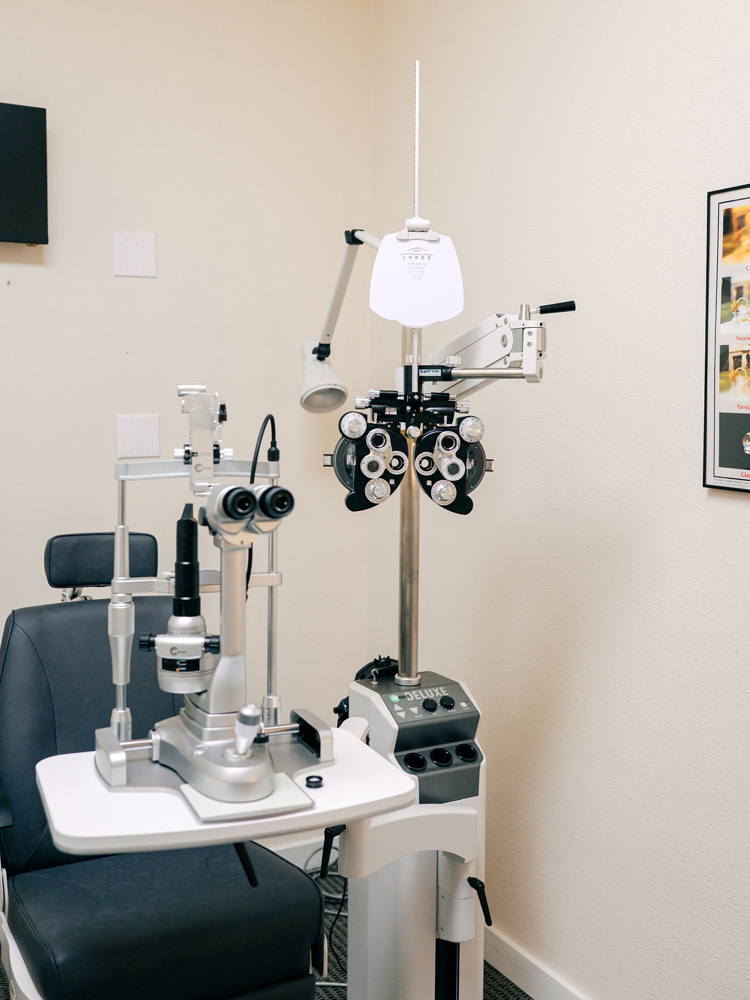 Optometry examination room with slit lamp, phoropter, and ophthalmic equipment, a black chair, a framed poster on the wall.