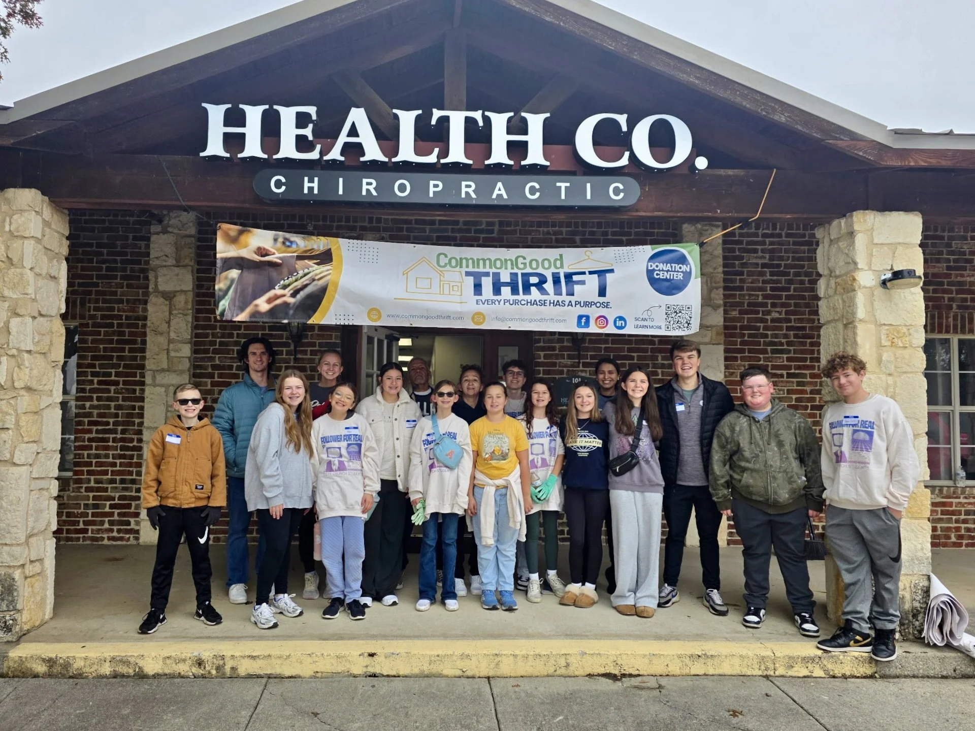 Group of young people and adults standing in front of a brick building with a sign that reads 'HEALTH CO. CHIROPRACTIC' and a banner promoting a thrift donation center, smiling for a group photo.