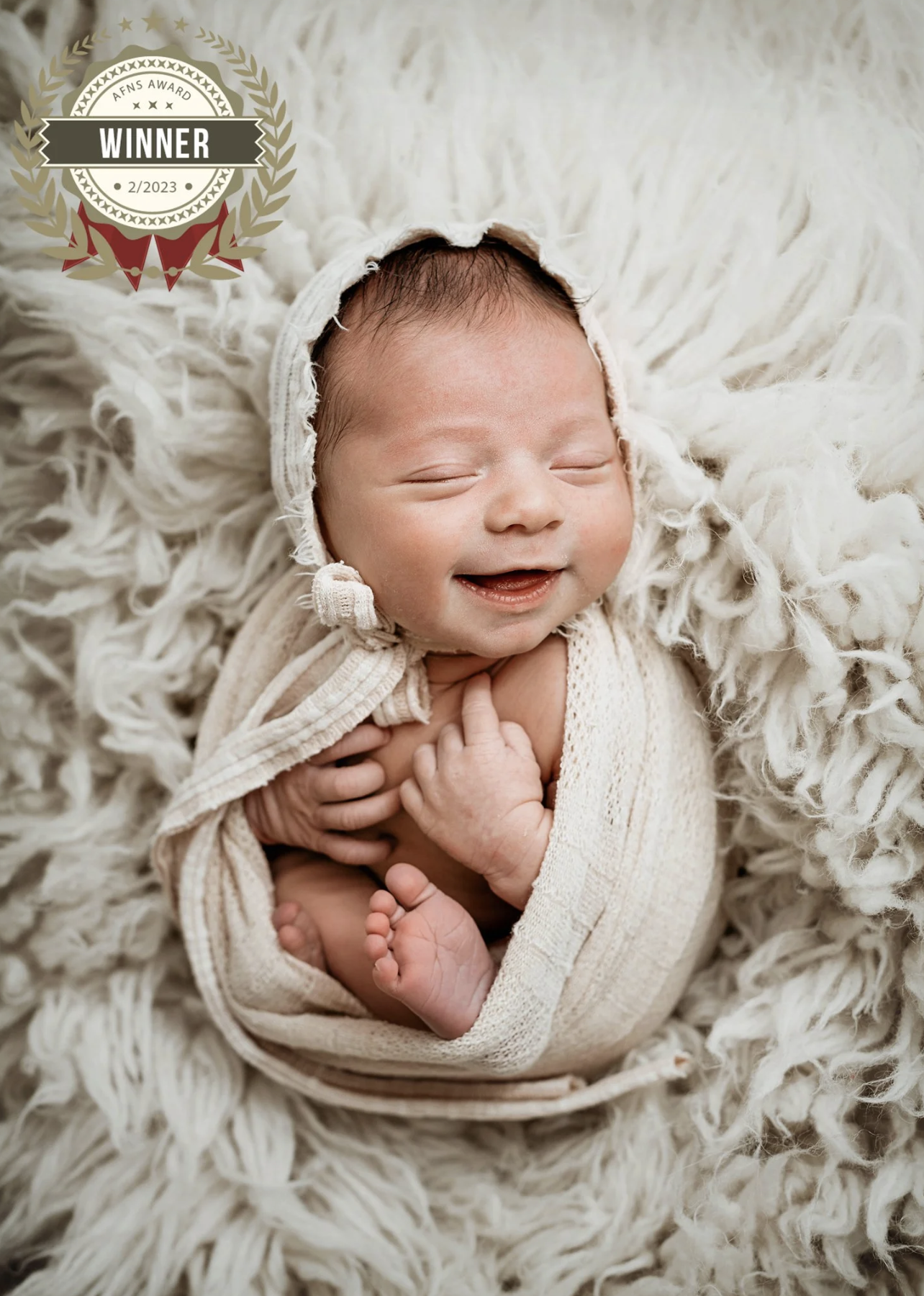 A smiling baby wrapped in beige fabric, resting on a fluffy white blanket with a sun hat, on a soft textured surface, with a "Winner" award badge on the top left corner.