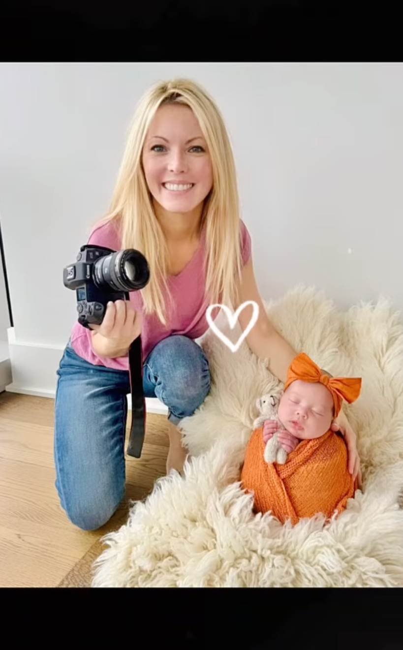 A smiling woman holding a camera kneels next to a newborn baby wrapped in an orange blanket and wearing an orange bow, sitting on a fluffy cream-colored rug with a stuffed animal.
