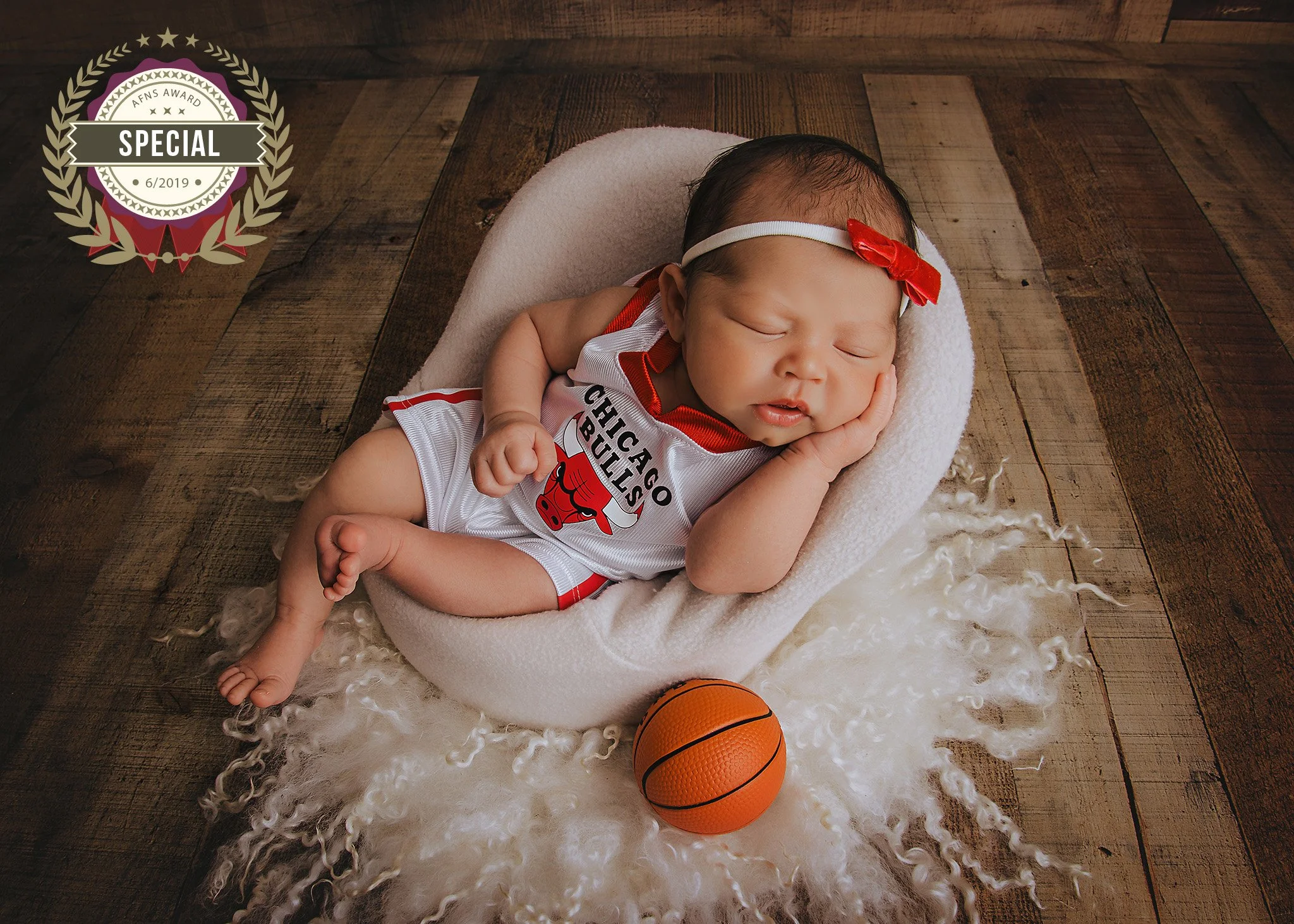 A sleeping baby wearing a Chicago Bulls basketball jersey and a white headband with a red bow, lying on a soft cream-colored blanket on a wooden floor, with a small orange basketball nearby.