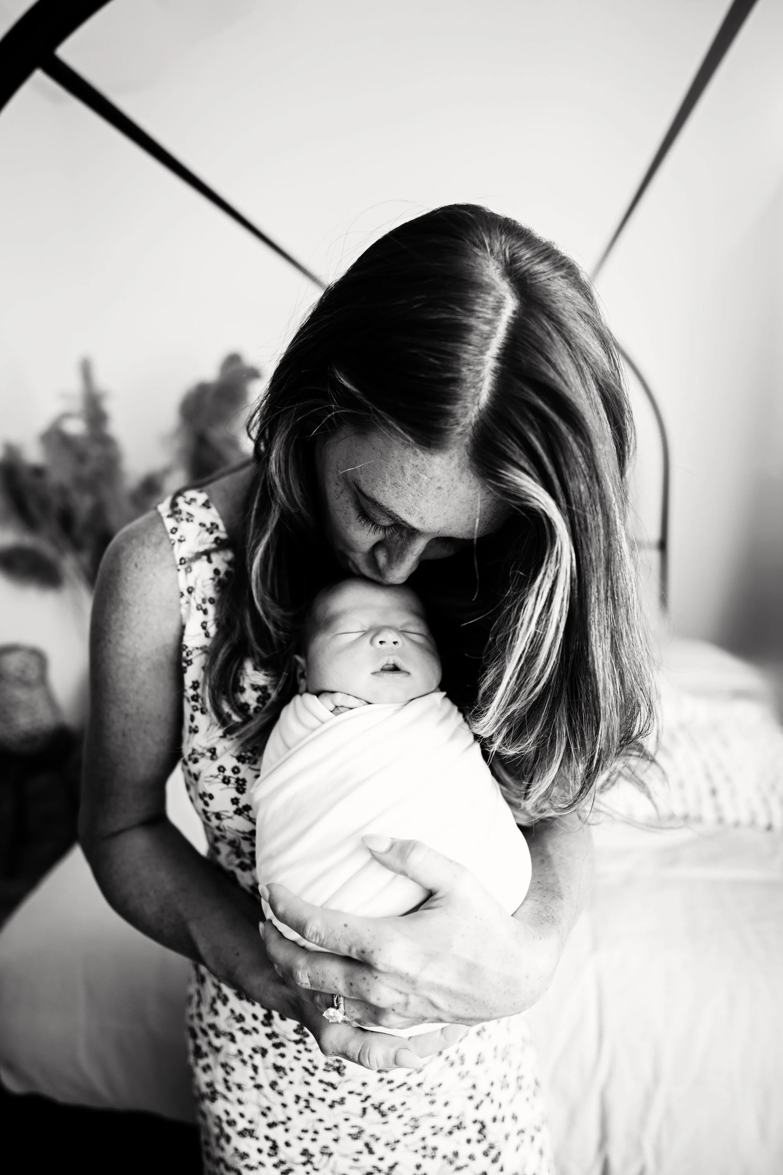 Black and white photograph of a woman holding a newborn baby while leaning down to kiss the baby's head. The woman is wearing a patterned dress and has long hair.
