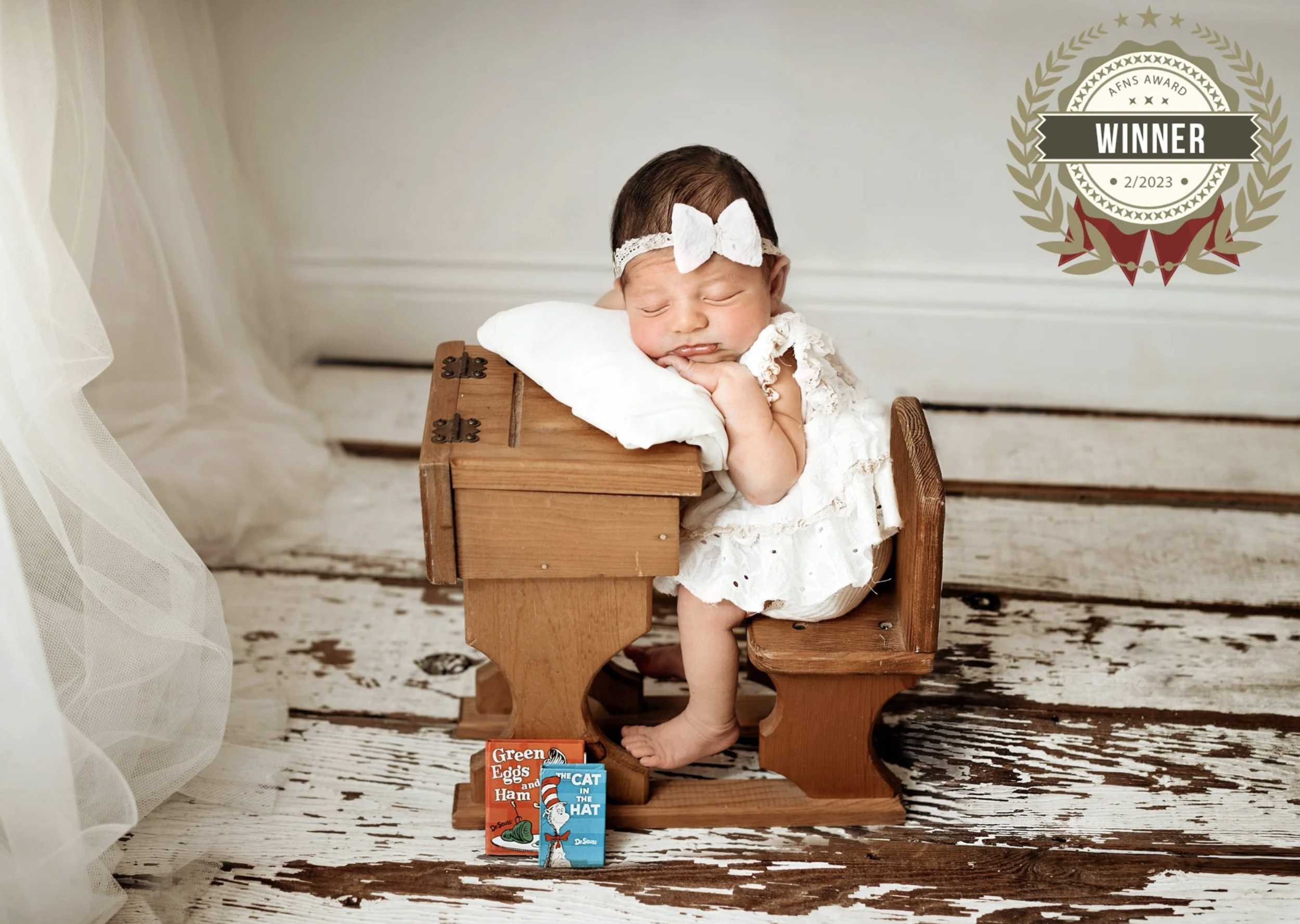 A sleeping baby with a lace headband and white dress, resting on a small wooden bench, with Dr. Seuss books at her feet, on a rustic wooden floor with white walls.