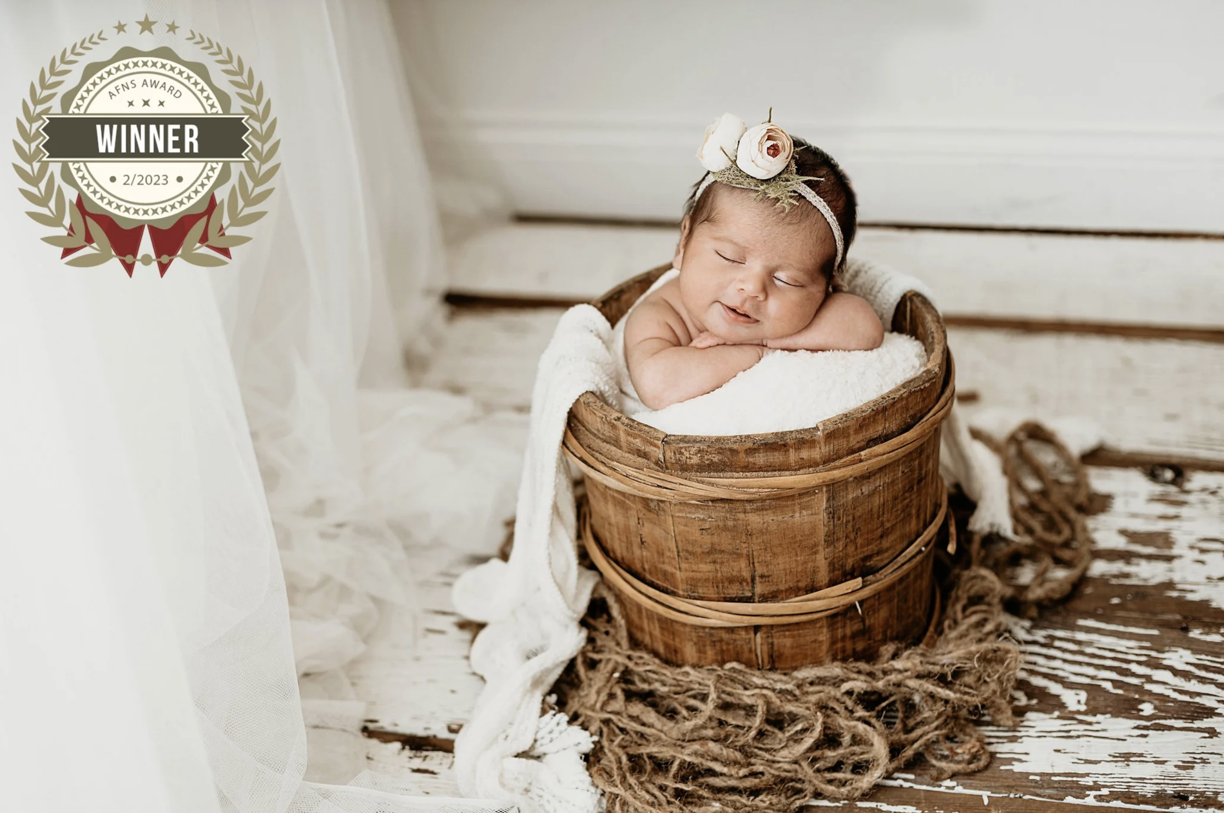 A newborn baby is resting in a wooden basket, wearing a floral headband, with a white blanket. The background includes a rustic wooden floor and white backdrop with a victory award badge on the left side.