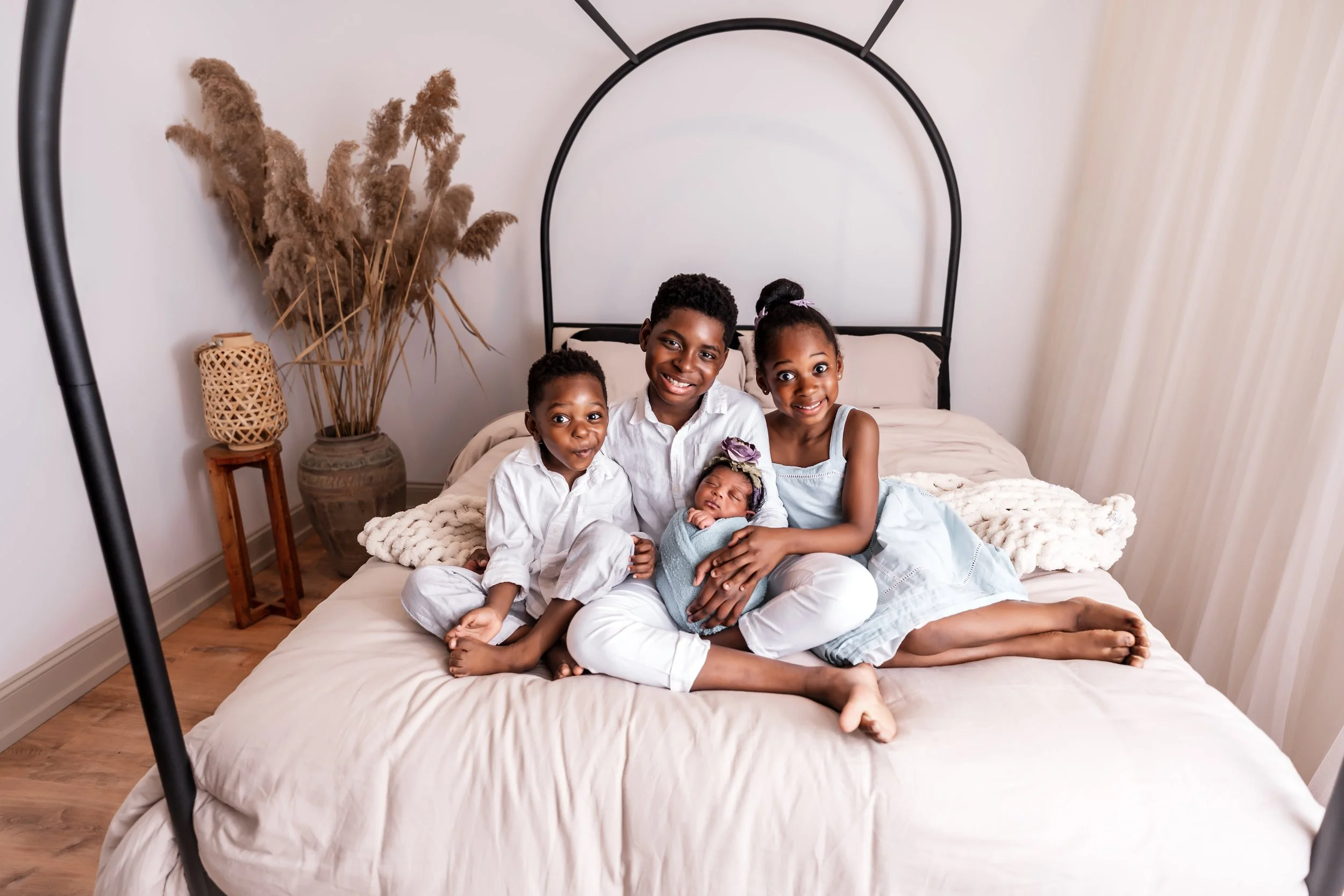 A family of five sitting on a bed, smiling at the camera in a bedroom with beige walls, wooden flooring, and a large potted plant with dried pampas grass next to it.