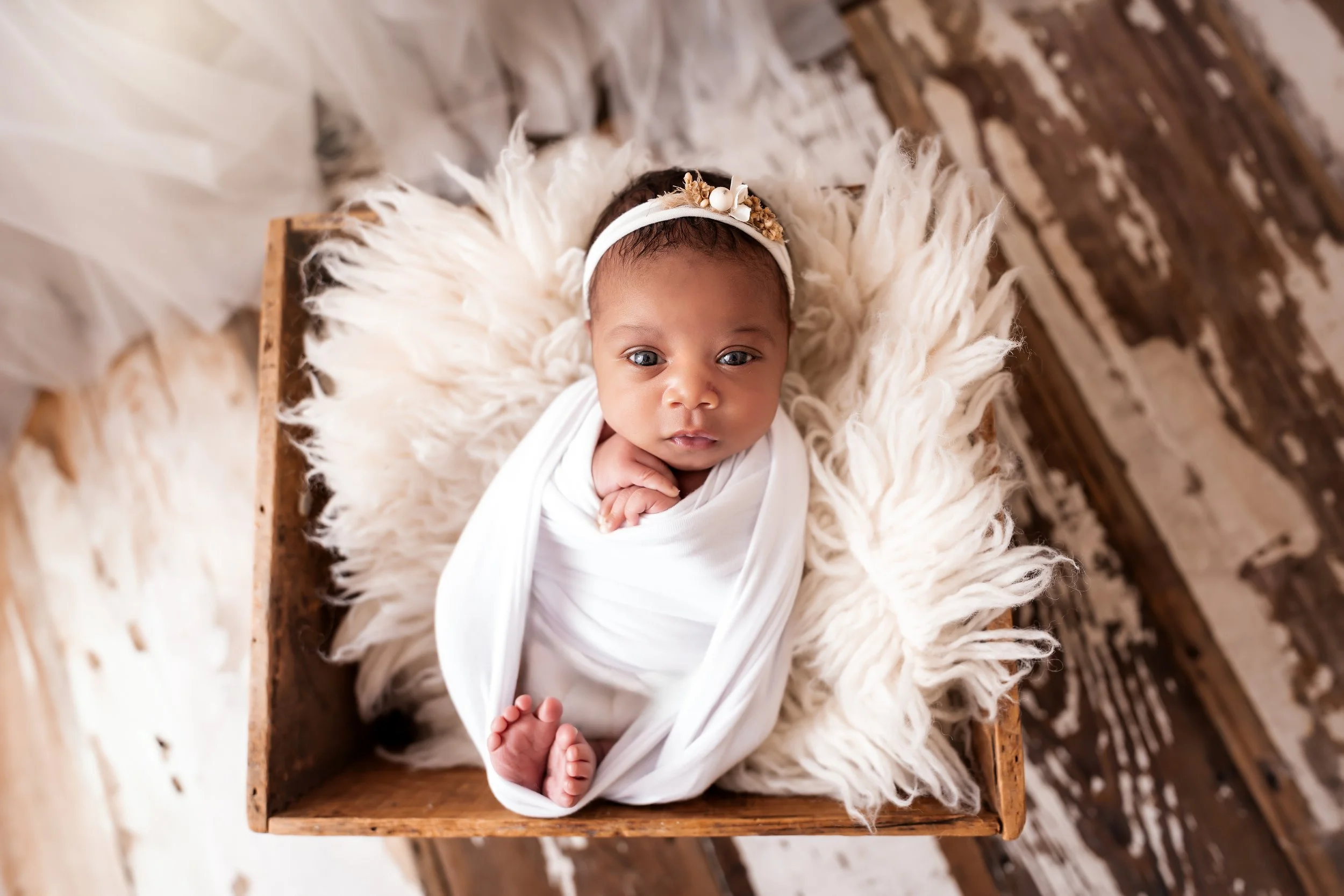 A baby with dark hair and bright blue eyes lying on a fluffy cream-colored blanket inside a rustic wooden box, wearing a headband with flowers and a white outfit, on a wooden surface.