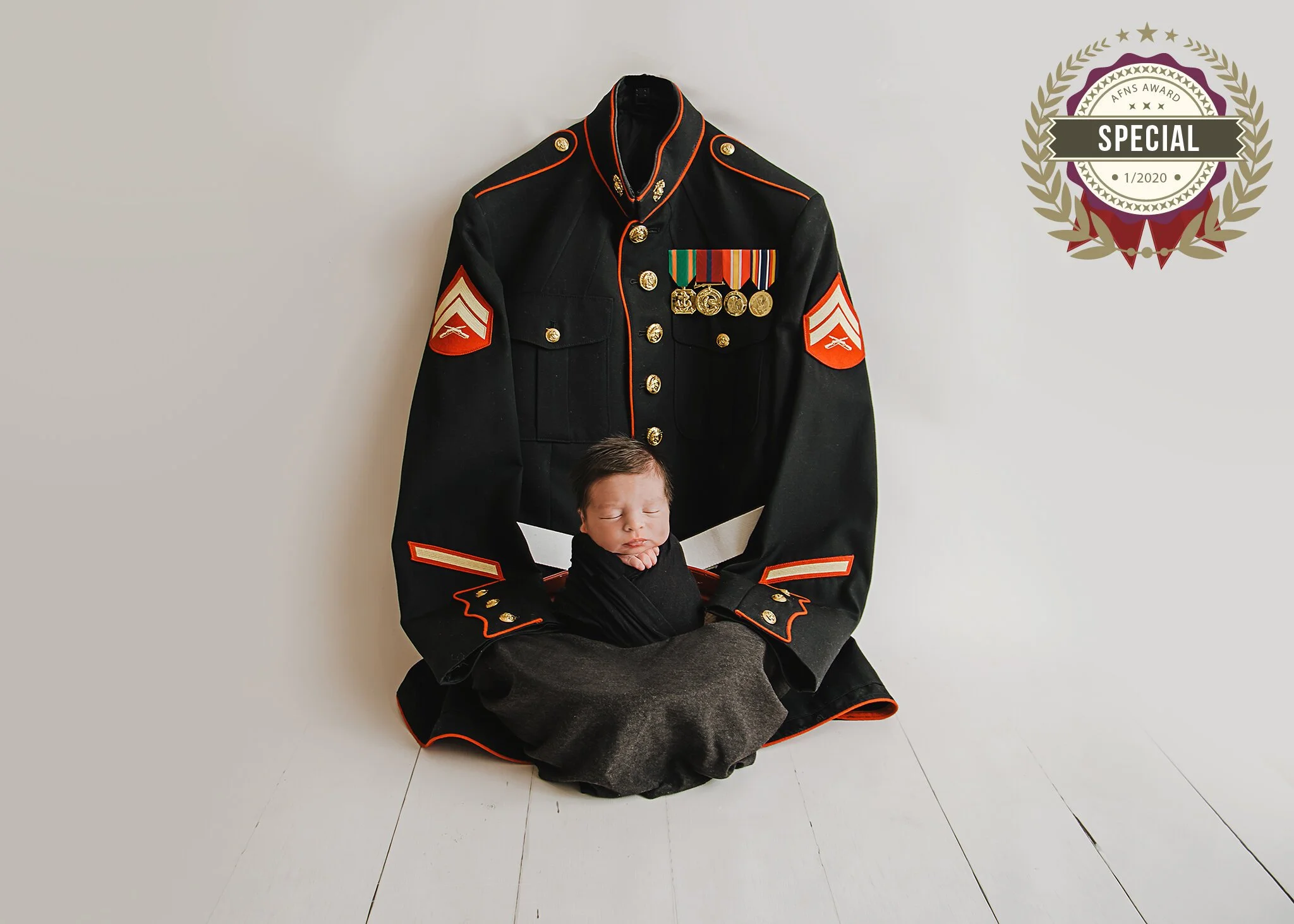 A person dressed in a vintage military dress uniform sitting on the floor, with a large military jacket hanging on the wall behind them. The jacket is decorated with medals and insignia, and the person has their eyes closed with their hands resting on their lap.