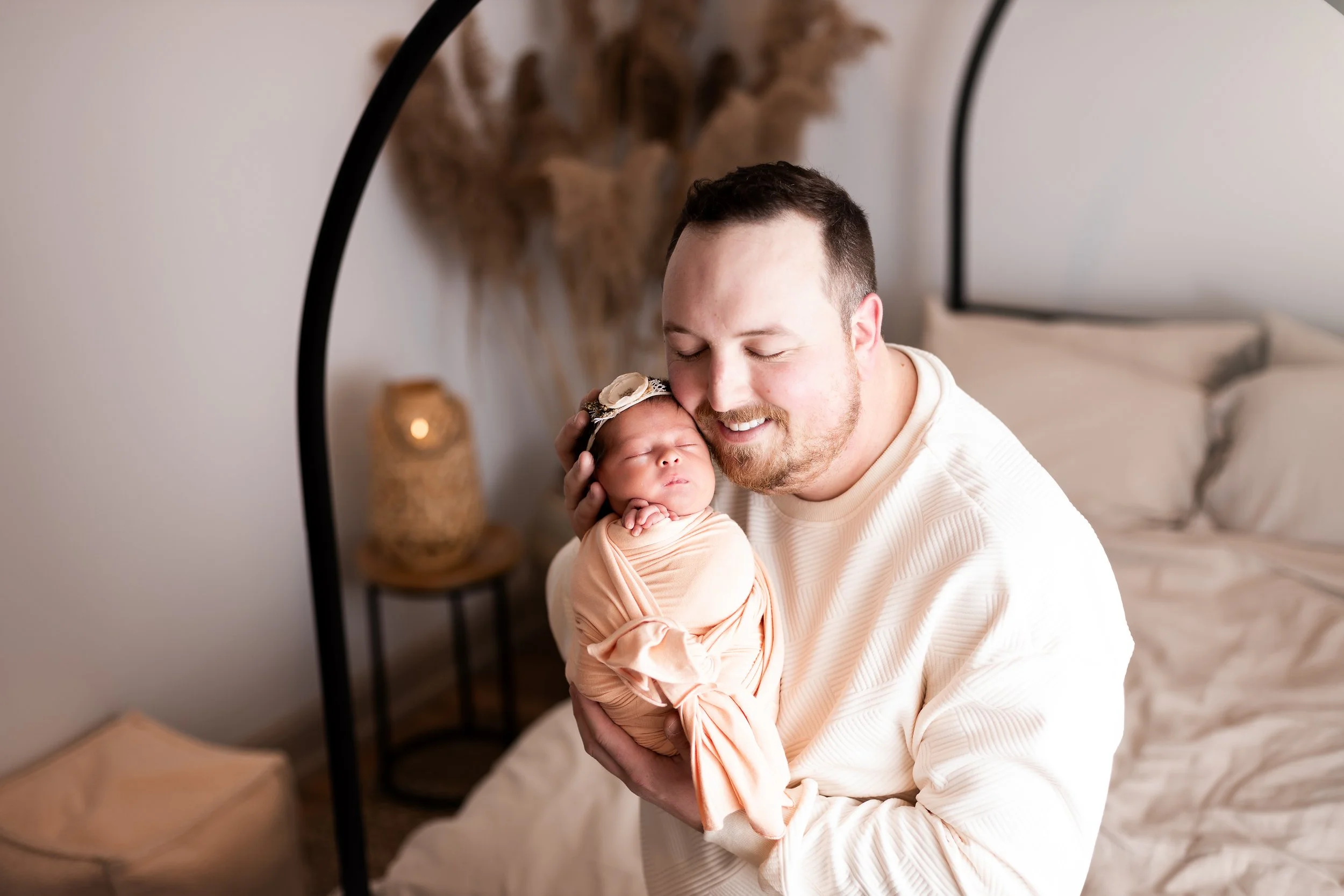 A man with short dark hair and a beard smiling while holding a sleeping baby with a floral headband. They are indoors in a cozy room with neutral decor and warm lighting.