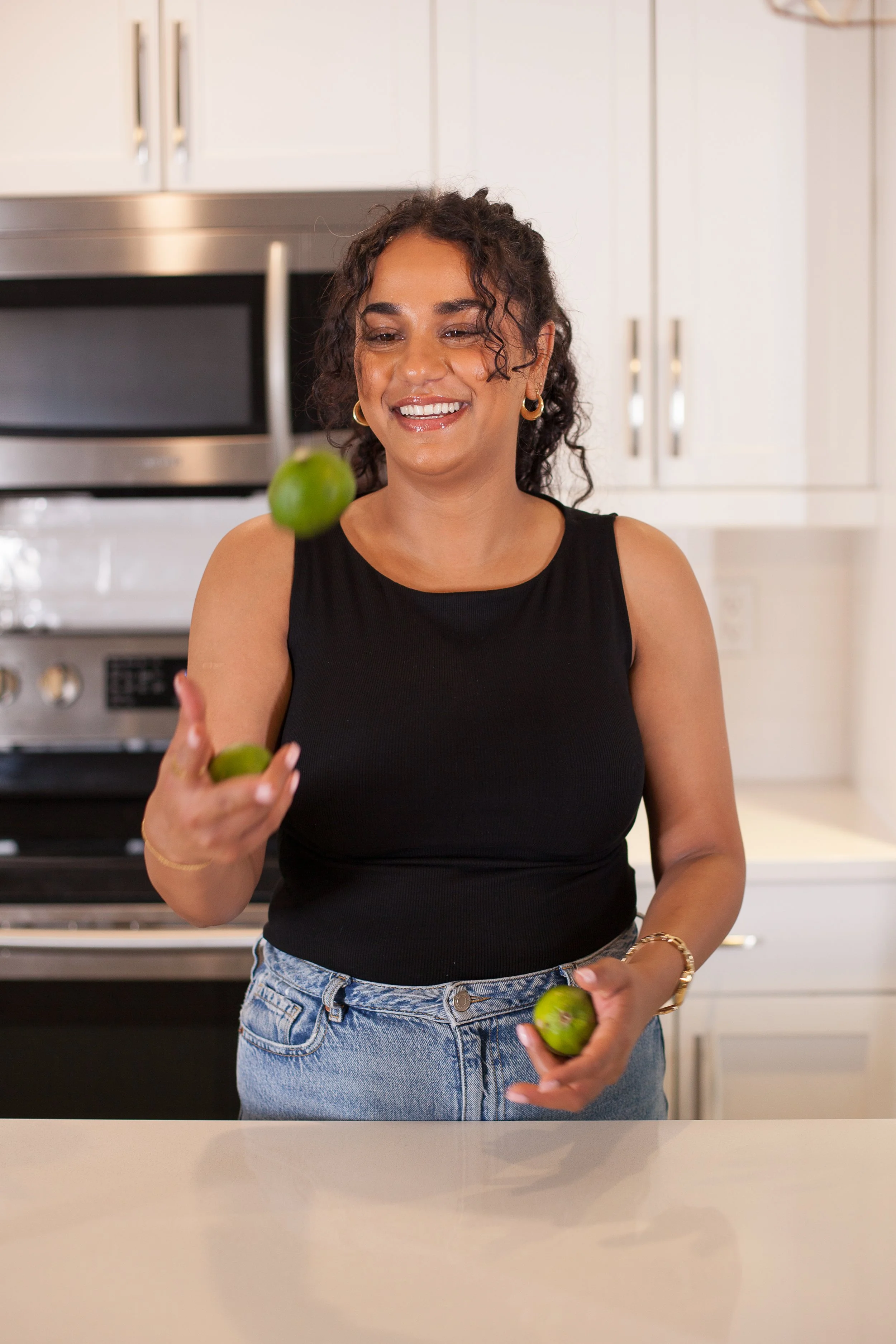 A woman in a black tank top and jeans is in a kitchen tossing green limes in the air, smiling and looking playful.