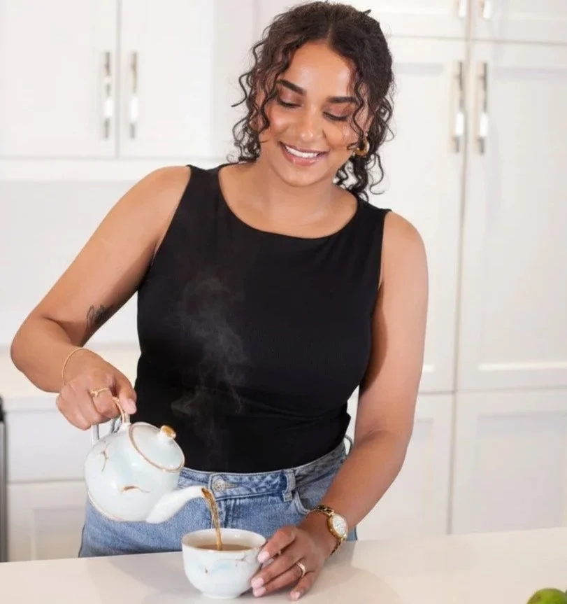 A woman in a black sleeveless top and blue jeans pours tea from a teapot into a cup in a kitchen.