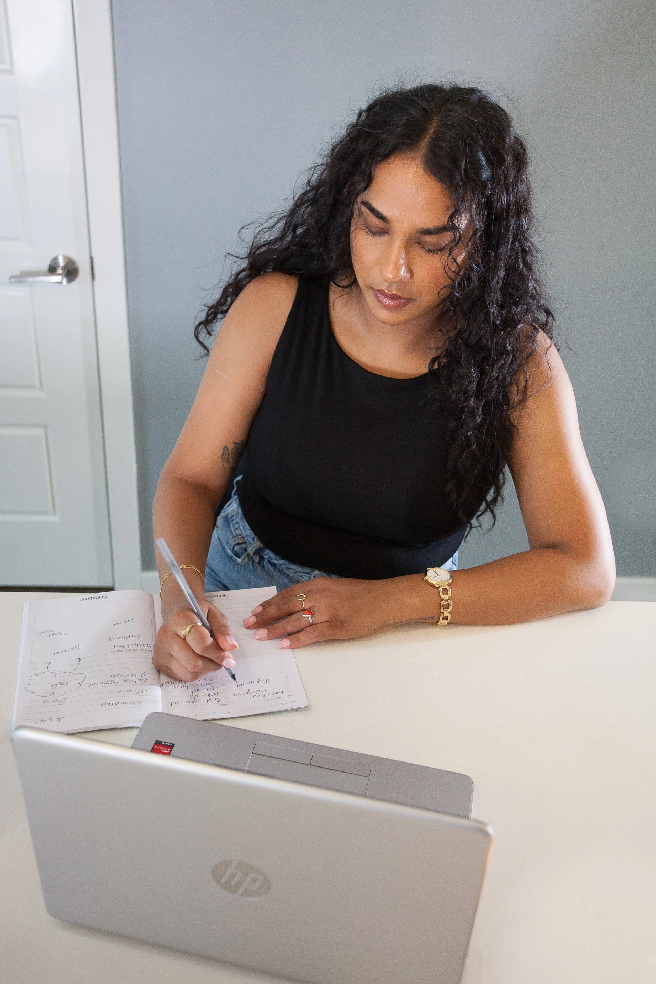 A woman with long curly black hair, wearing a black sleeveless top and jewelry, is sitting at a desk writing in a notebook. There is a laptop in front of her and a grey wall in the background.