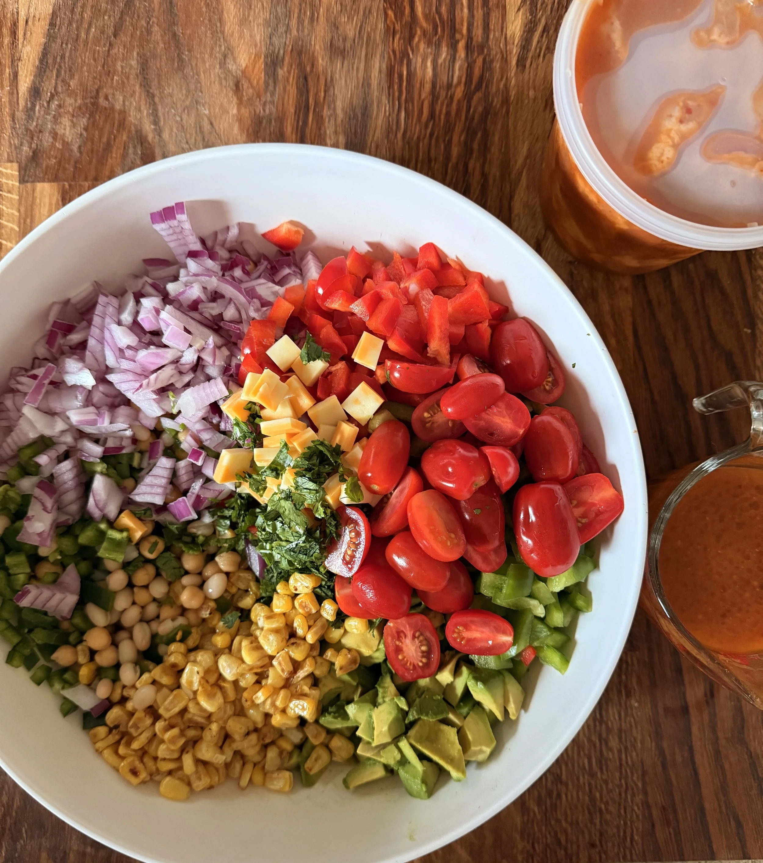 A white bowl filled with chopped red onions, red cherry tomatoes, diced yellow cheese, chopped red bell peppers, chopped celery, chopped fresh herbs, and cooked chickpeas. A container of soup or sauce is partially visible in the upper right corner, next to the bowl, on a wooden surface.