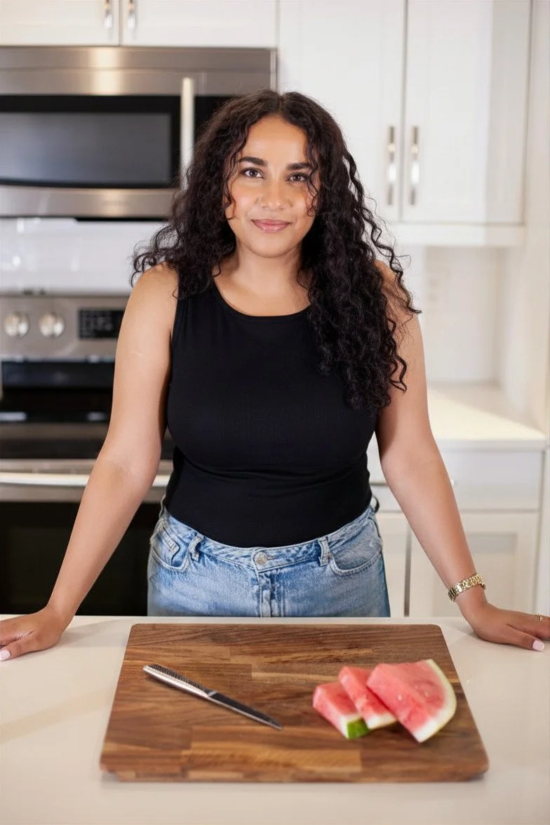 Woman with curly hair in a black sleeveless top and blue jeans standing behind a kitchen counter with a wooden cutting board, a black knife, and slices of watermelon.