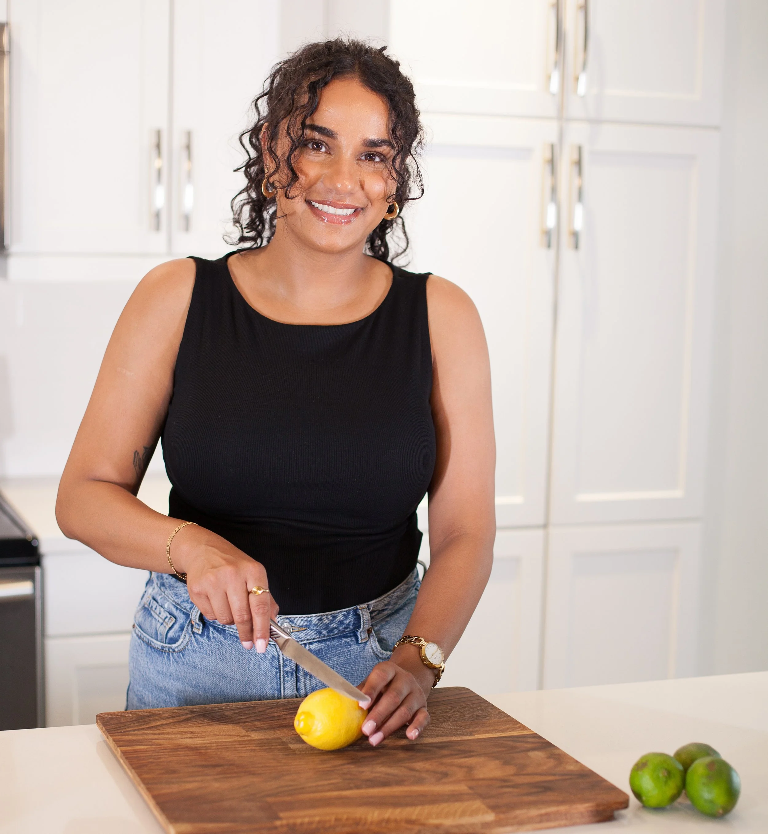 A woman with curly dark hair, wearing a black sleeveless top, is slicing a yellow lemon on a wooden cutting board in a bright kitchen. There are green limes in the foreground.