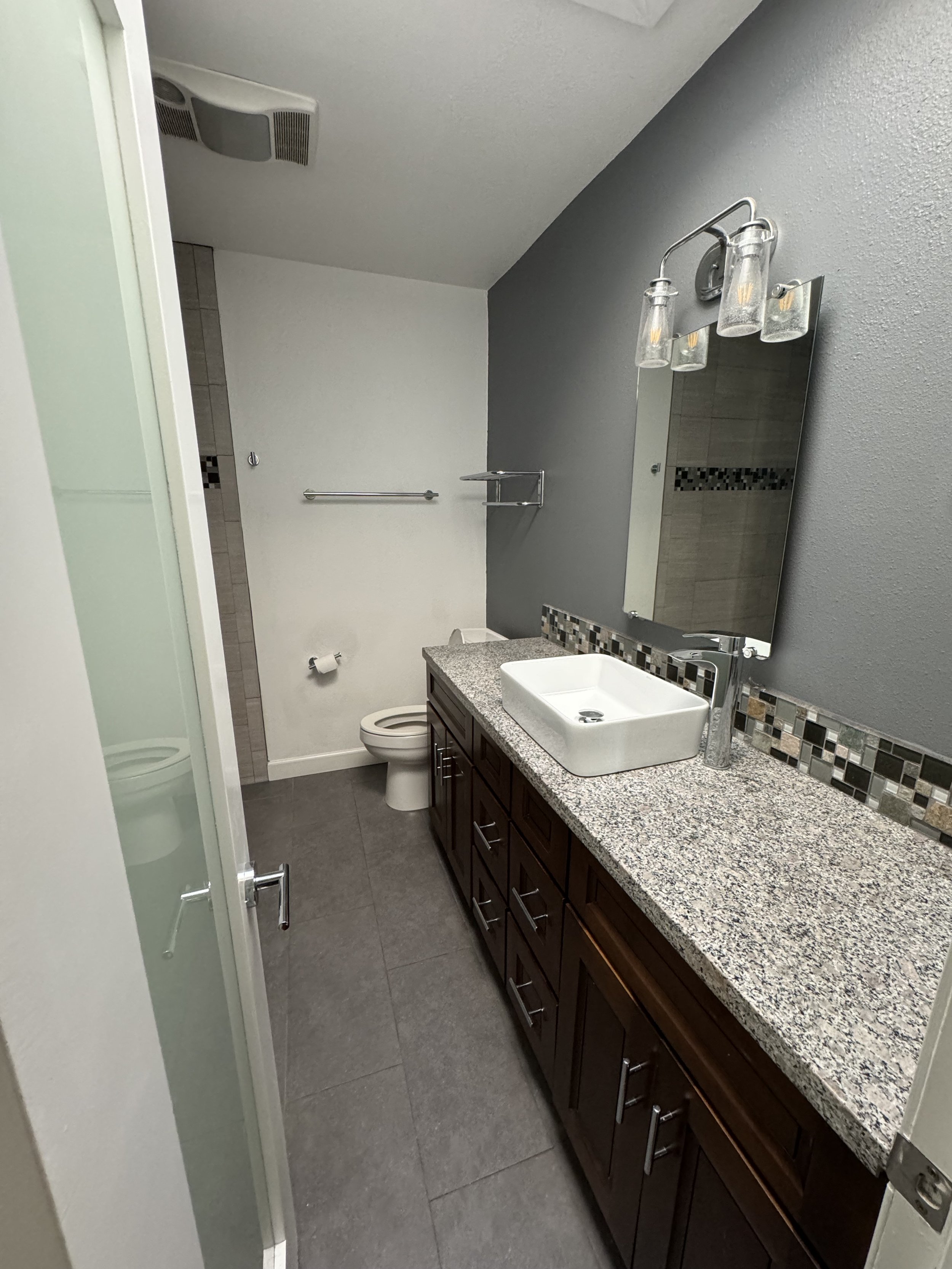 A modern bathroom with a granite countertop vanity, a white vessel sink, a large mirror, wall-mounted light fixtures, a toilet, a towel bar, and a shower stall with a frosted glass door.