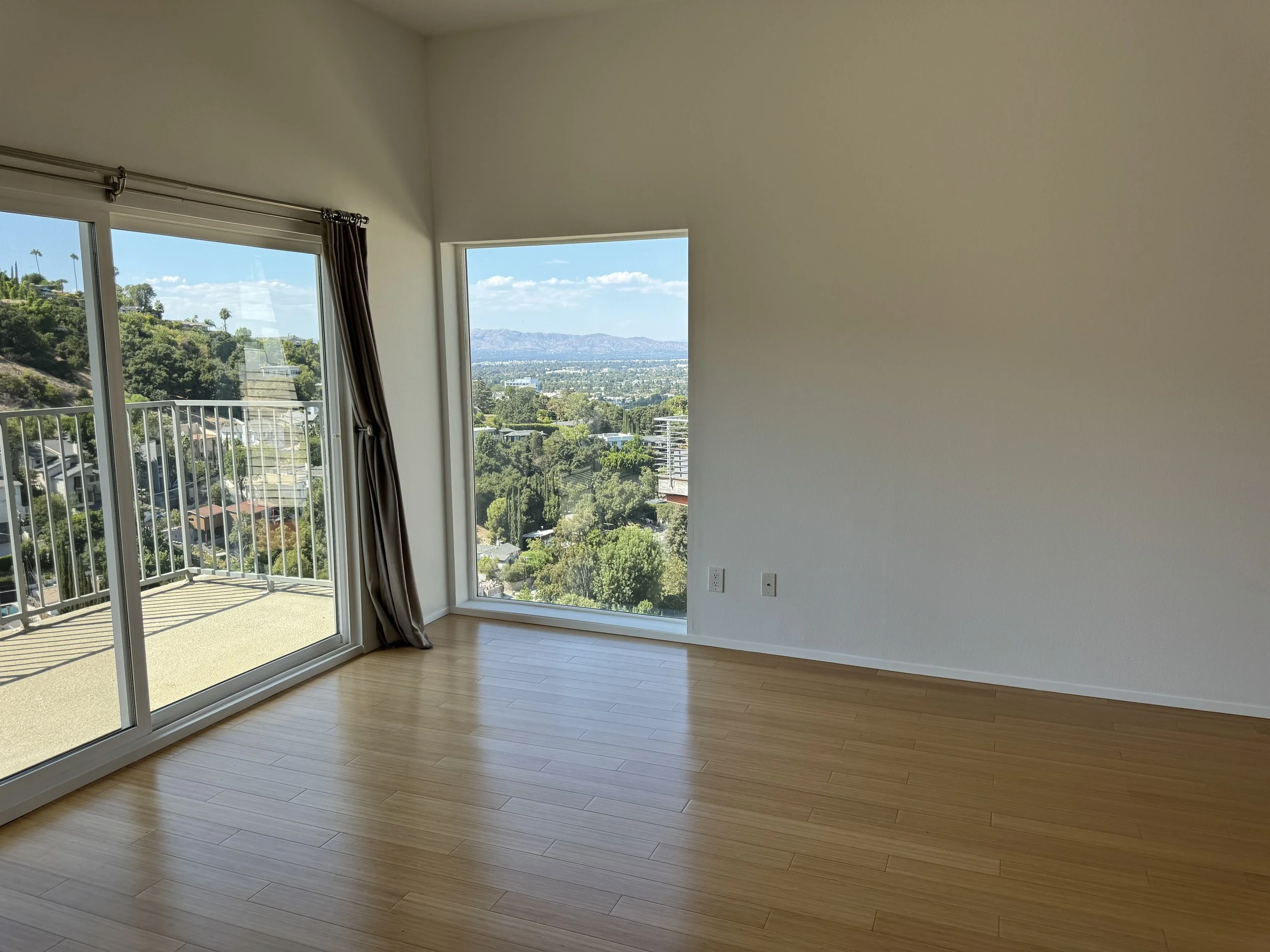Empty room with large windows showing a view of a city and greenery, wooden floor, white walls, and sliding glass door leading to a balcony.