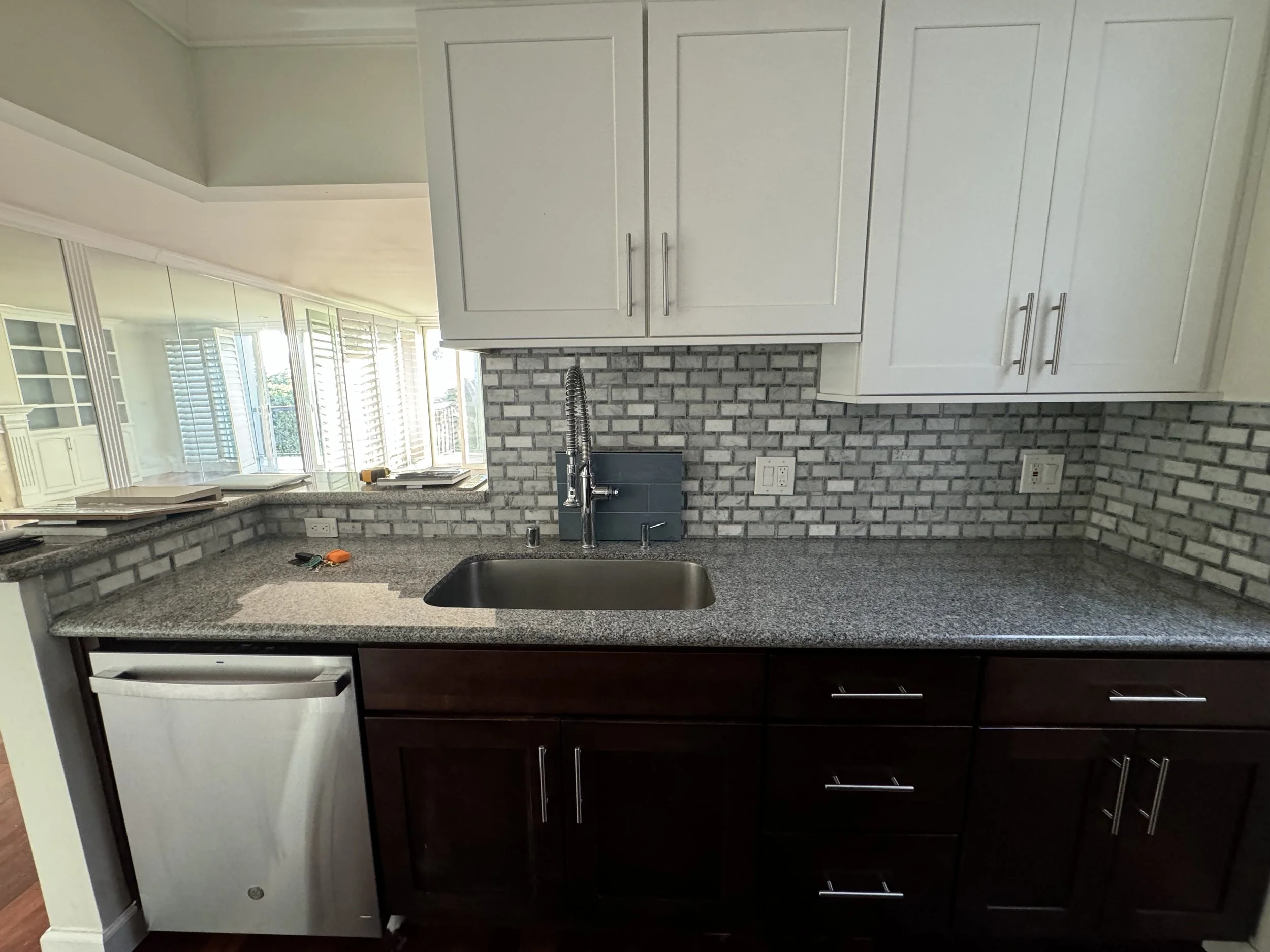 A kitchen with granite countertops, white upper cabinets, dark lower cabinets, brick-style backsplash, a stainless steel sink with a flexible hose faucet, and several tools and papers on the counter.