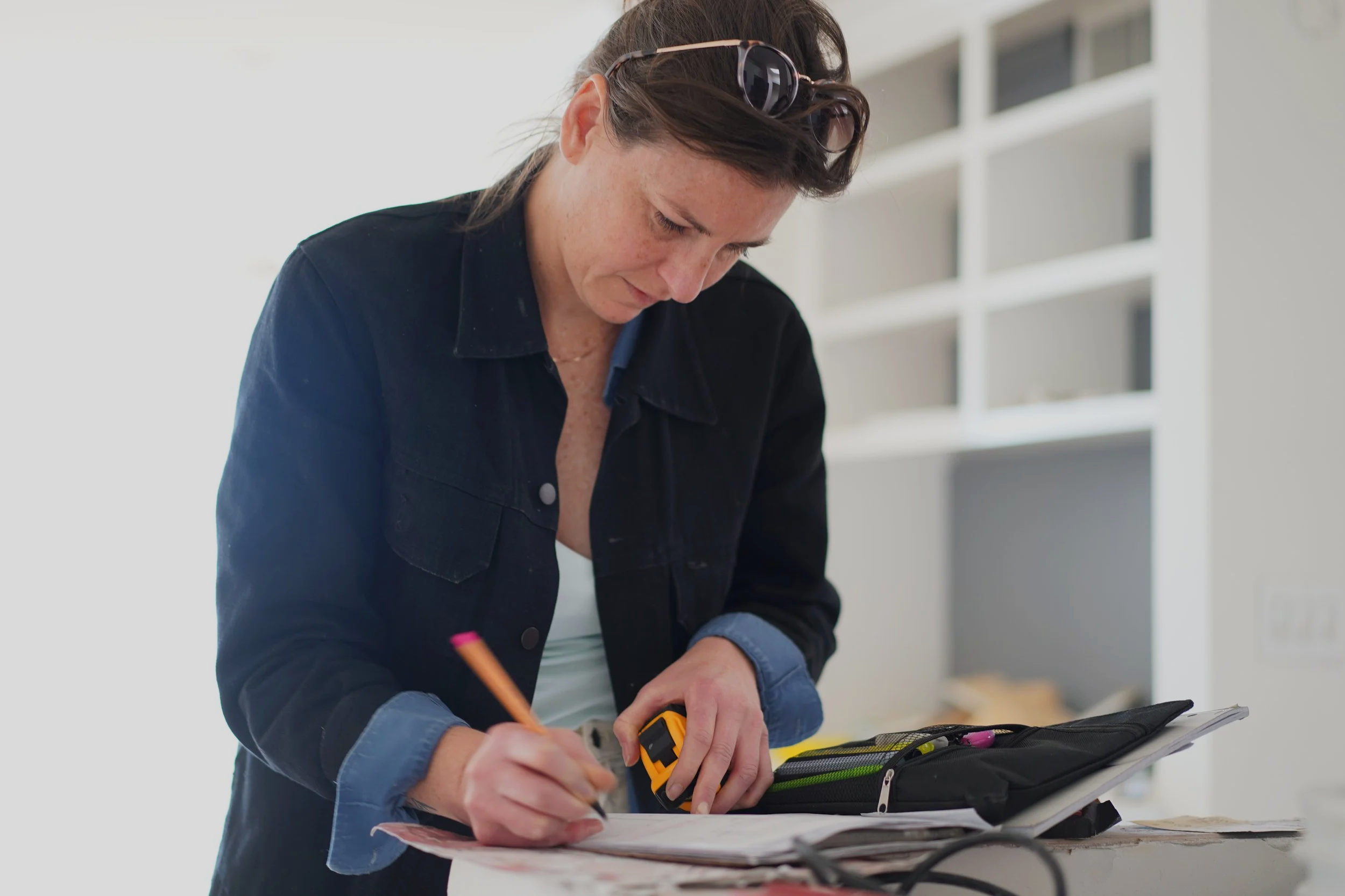 Woman working on a tablet with a pencil in hand, using a tape measure on a desk, with a toolbox nearby.