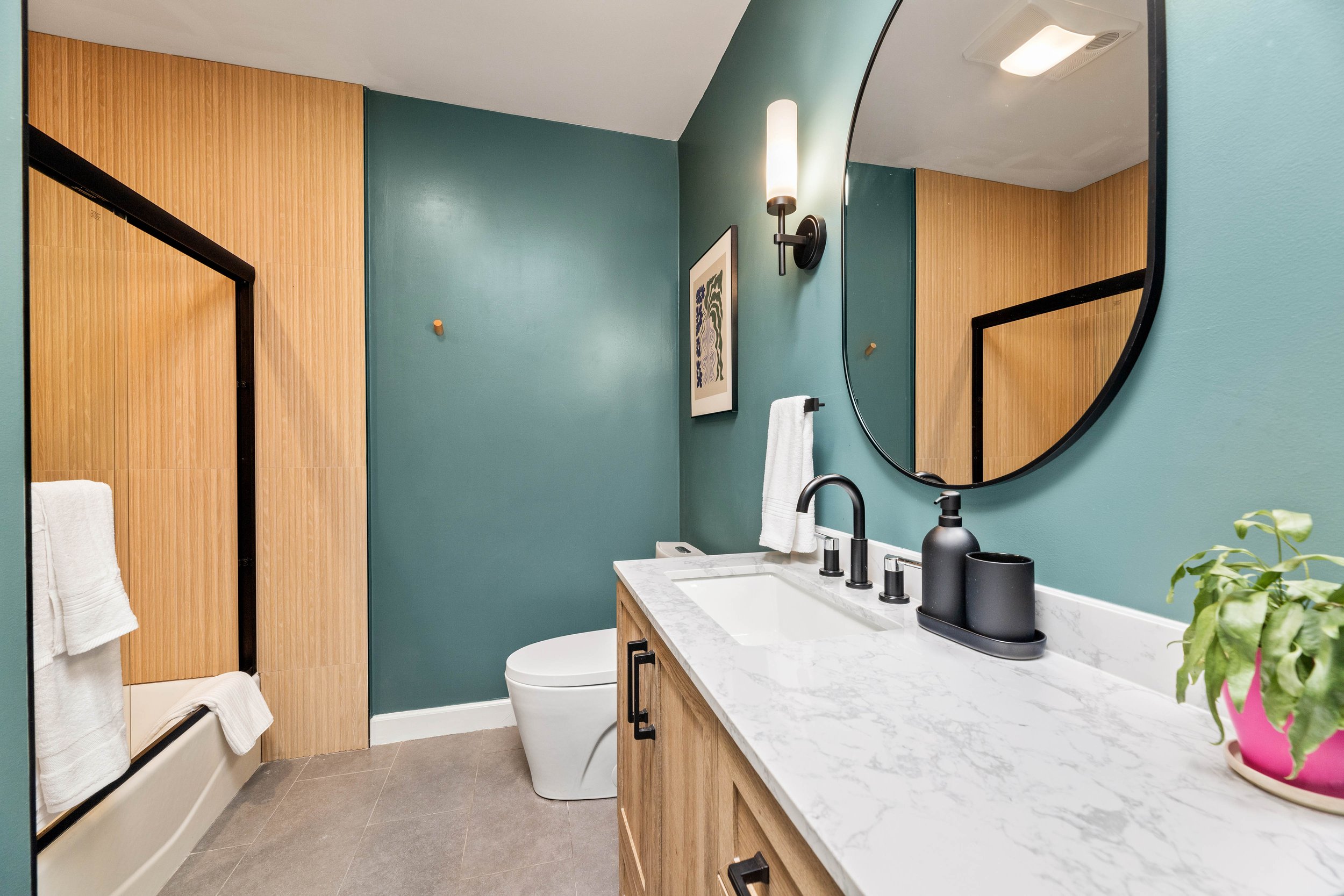 Bathroom with green wall, wooden paneling, marble countertop, black faucet, large round mirror, soap dispenser, potted plant, toilet, wall art, wall sconce, and towel rack.
