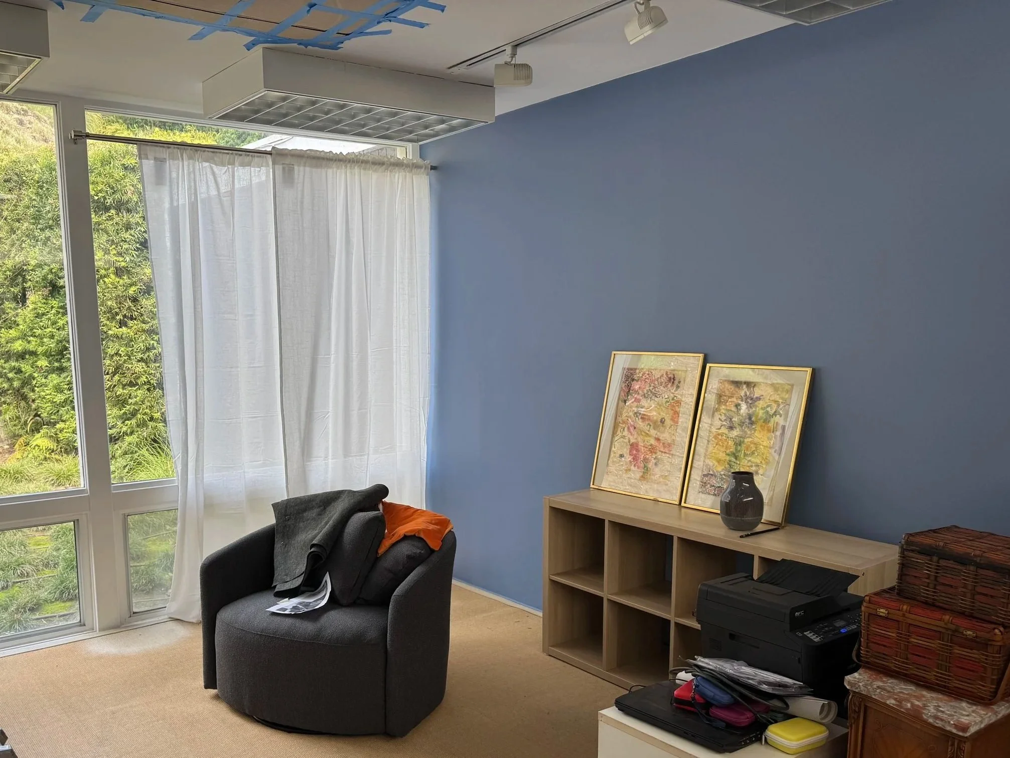 A cozy office space with a large window and white curtains, two framed artwork pieces leaning against a blue wall, a gray armchair with clothes on it, a wooden bookshelf, and a black printer on a white stand.
