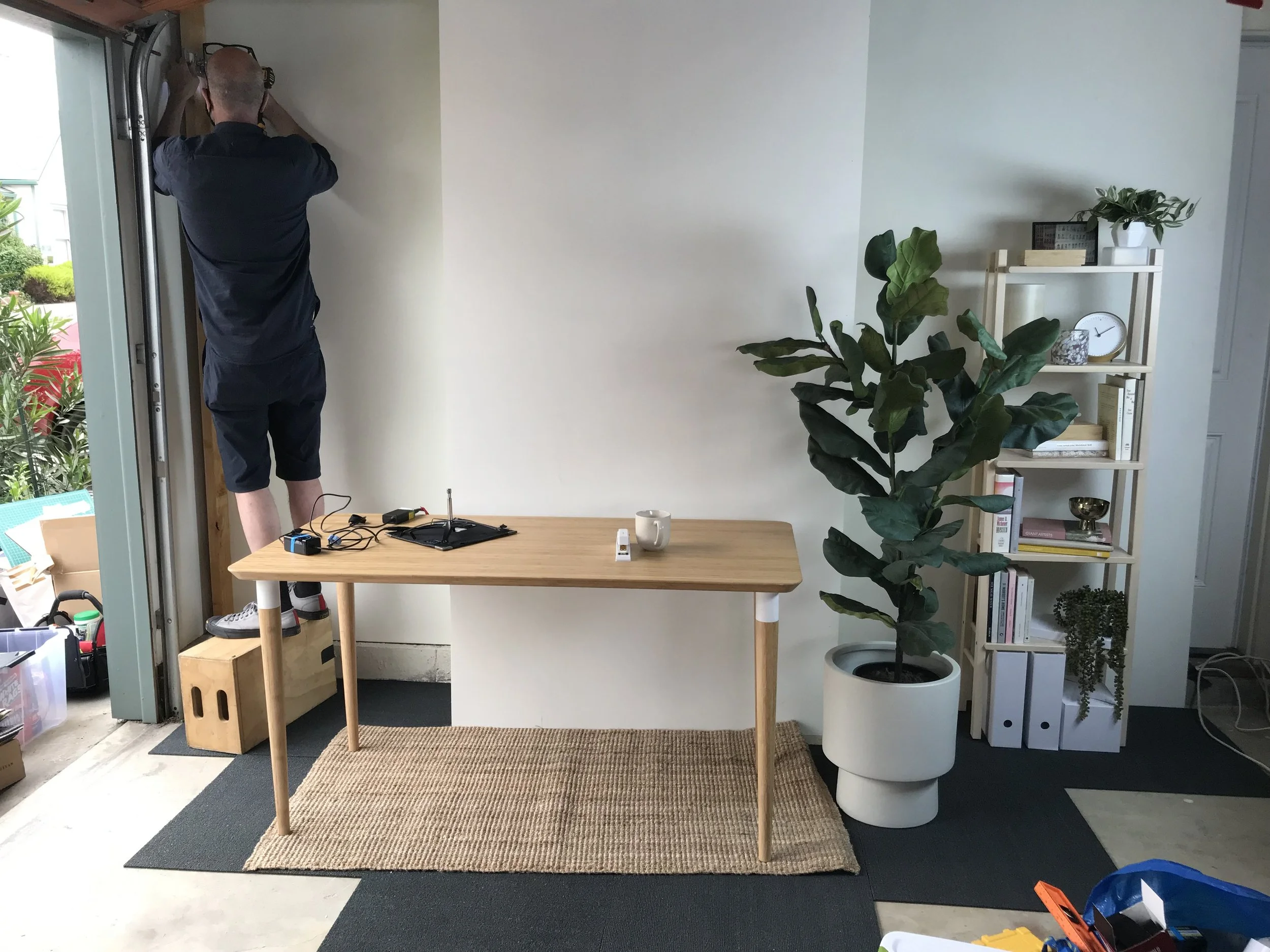A man with a shaved head wearing a black shirt and shorts is standing on a wooden table, working with tools near a doorway in a room. The room has a white wall, a large potted plant, and a white bookshelf with various decorative items and books.