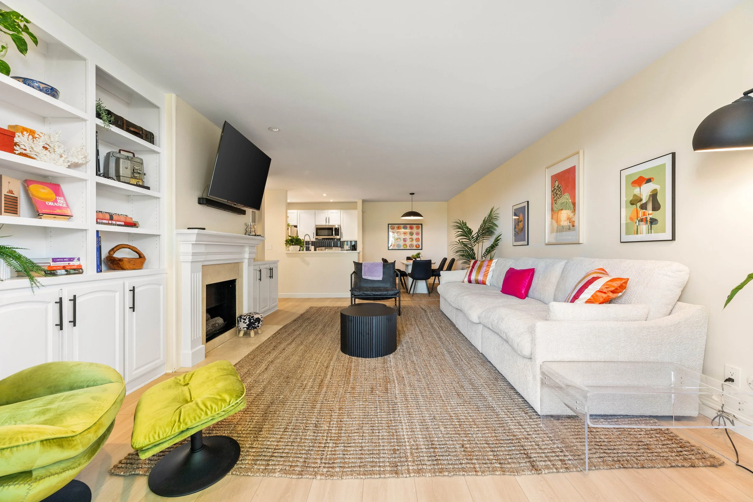 Bright living room with white sofa with colorful pillows, abstract framed art on the wall, potted plants, a black coffee table, chairs around a dining table, built-in shelves with books and decor, and a view of the kitchen area with white cabinets in the background.