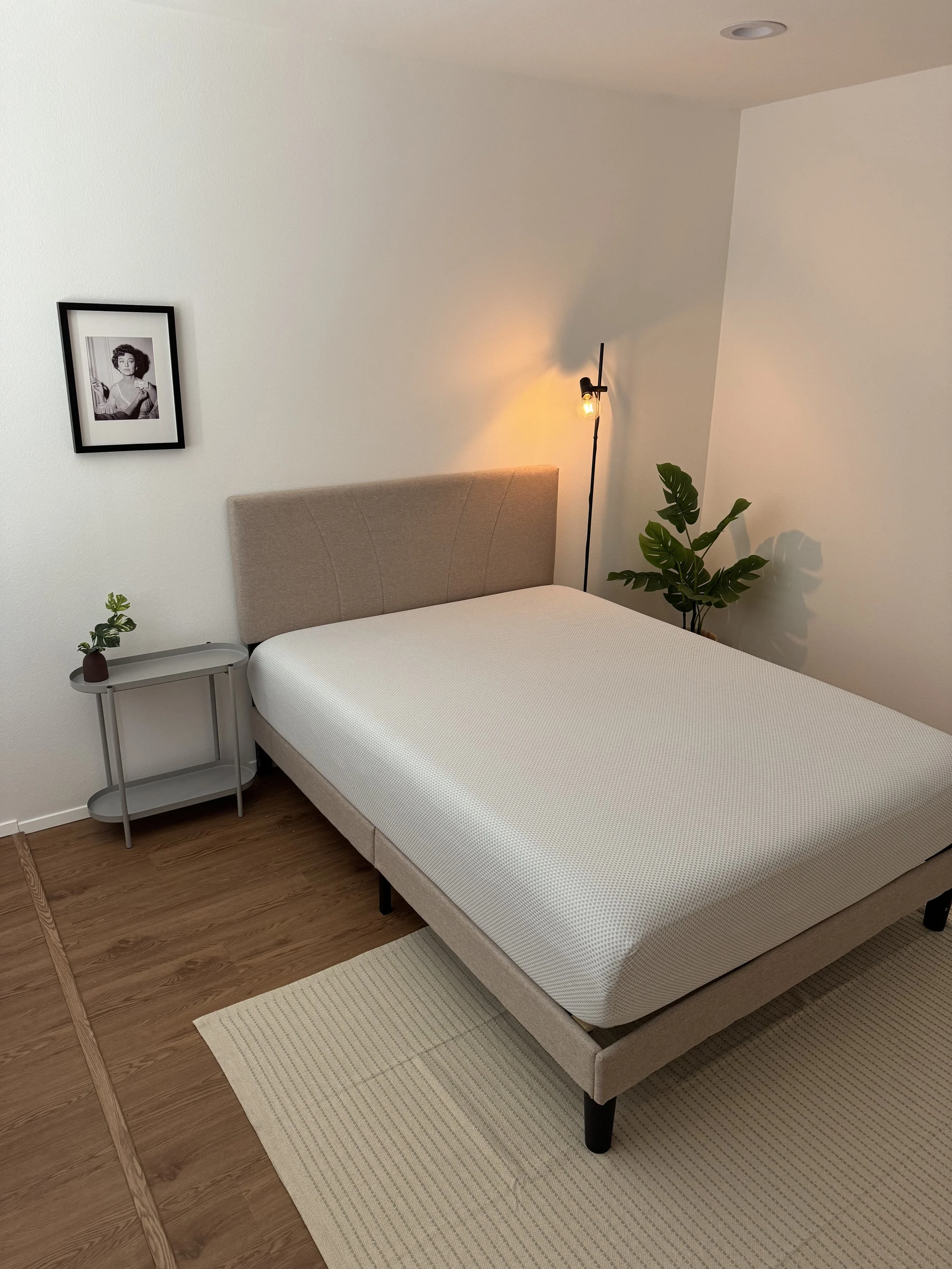 A bedroom with a beige upholstered bed, a beige headboard, a small side table with a potted plant, a framed black and white photograph of a woman, a floor lamp next to the bed, a plant with large green leaves, a wood floor, and a light-colored rug.