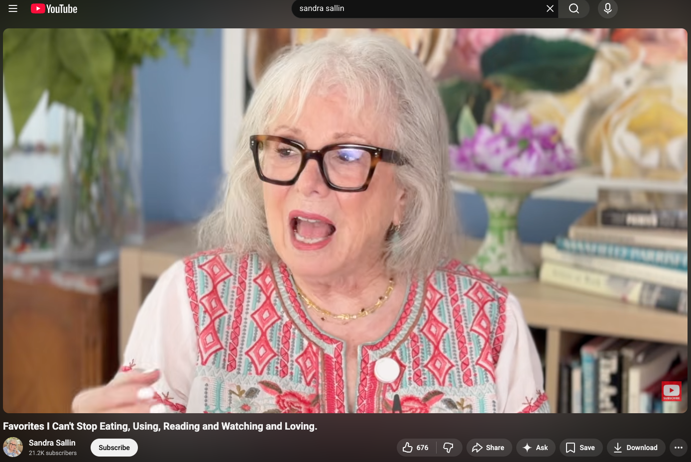 An older woman with white hair and glasses speaking during a video interview, sitting in front of a bookshelf and a decorative flower arrangement on a table.