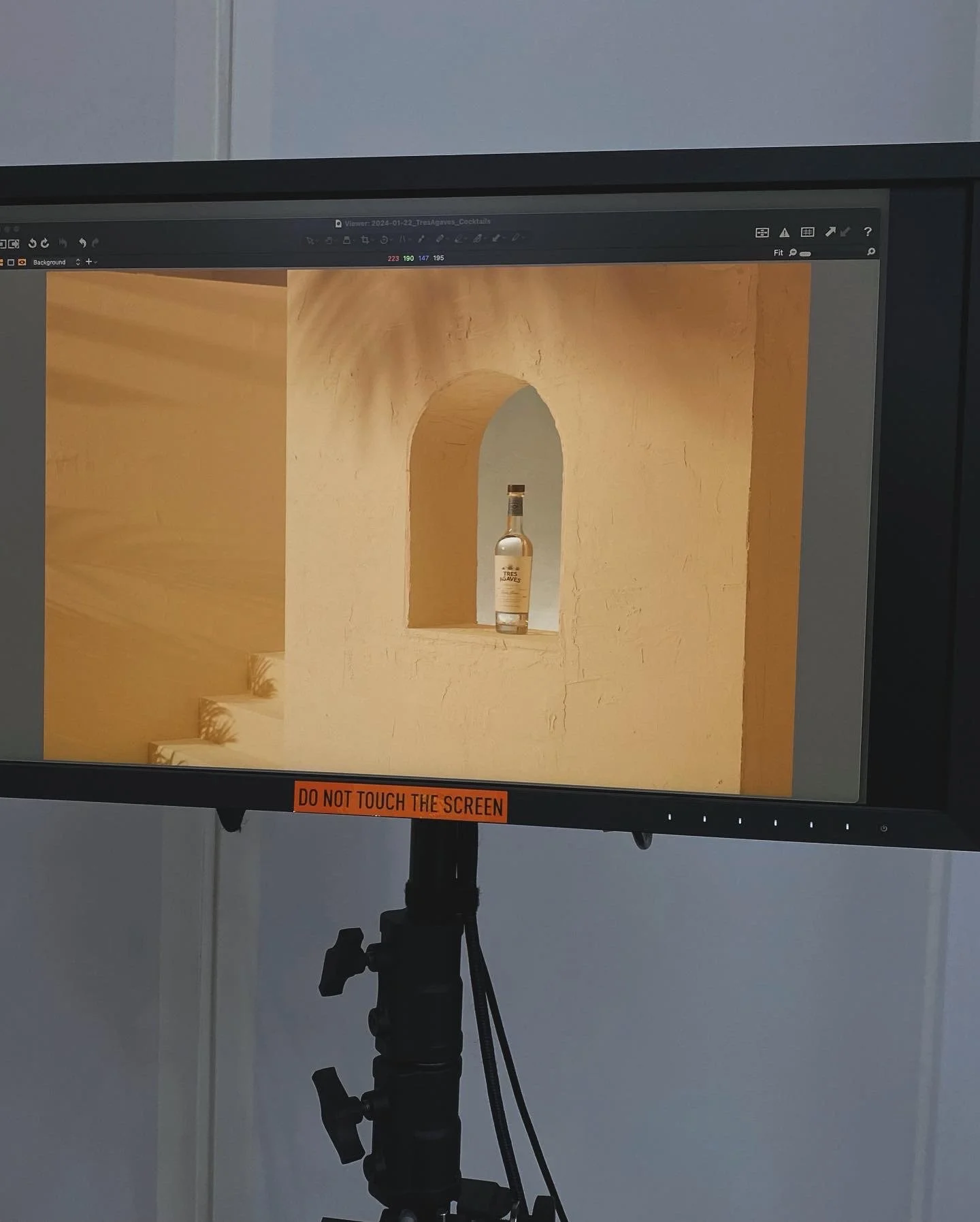 Photograph of a computer screen displaying an image of a bottle on a yellow textured wall with steps and an archway. A tripod is visible in front of the screen, with a sticker that reads 'DO NOT TOUCH THE SCREEN'.