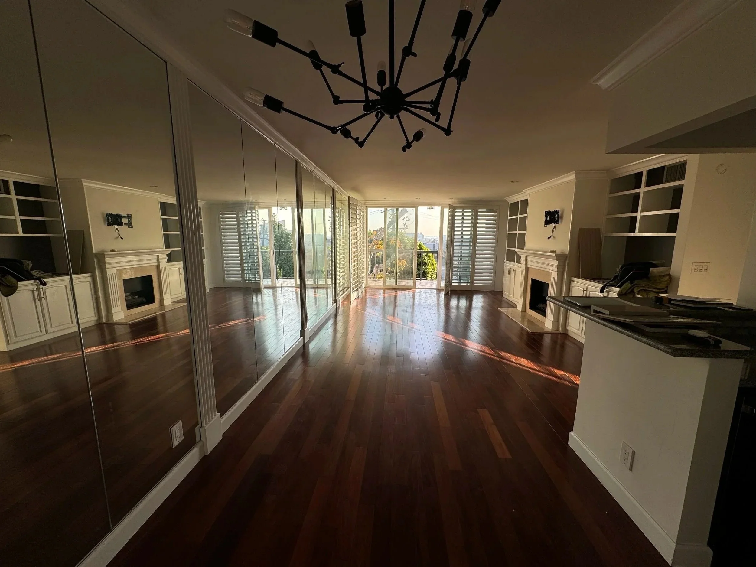 A spacious room with polished wooden floors, large glass sliding doors with white shutters, white built-in cabinets, and a black chandelier hanging from the ceiling. The room is brightly lit by natural sunlight.
