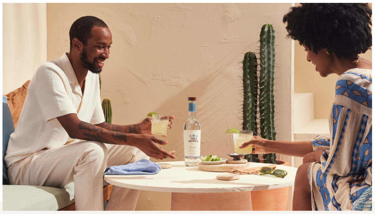 A man and woman sitting at a small round table outdoors, sharing drinks and smiling. The man is handing the woman a drink. There are cacti, a bottle of alcohol, and lime slices on the table.