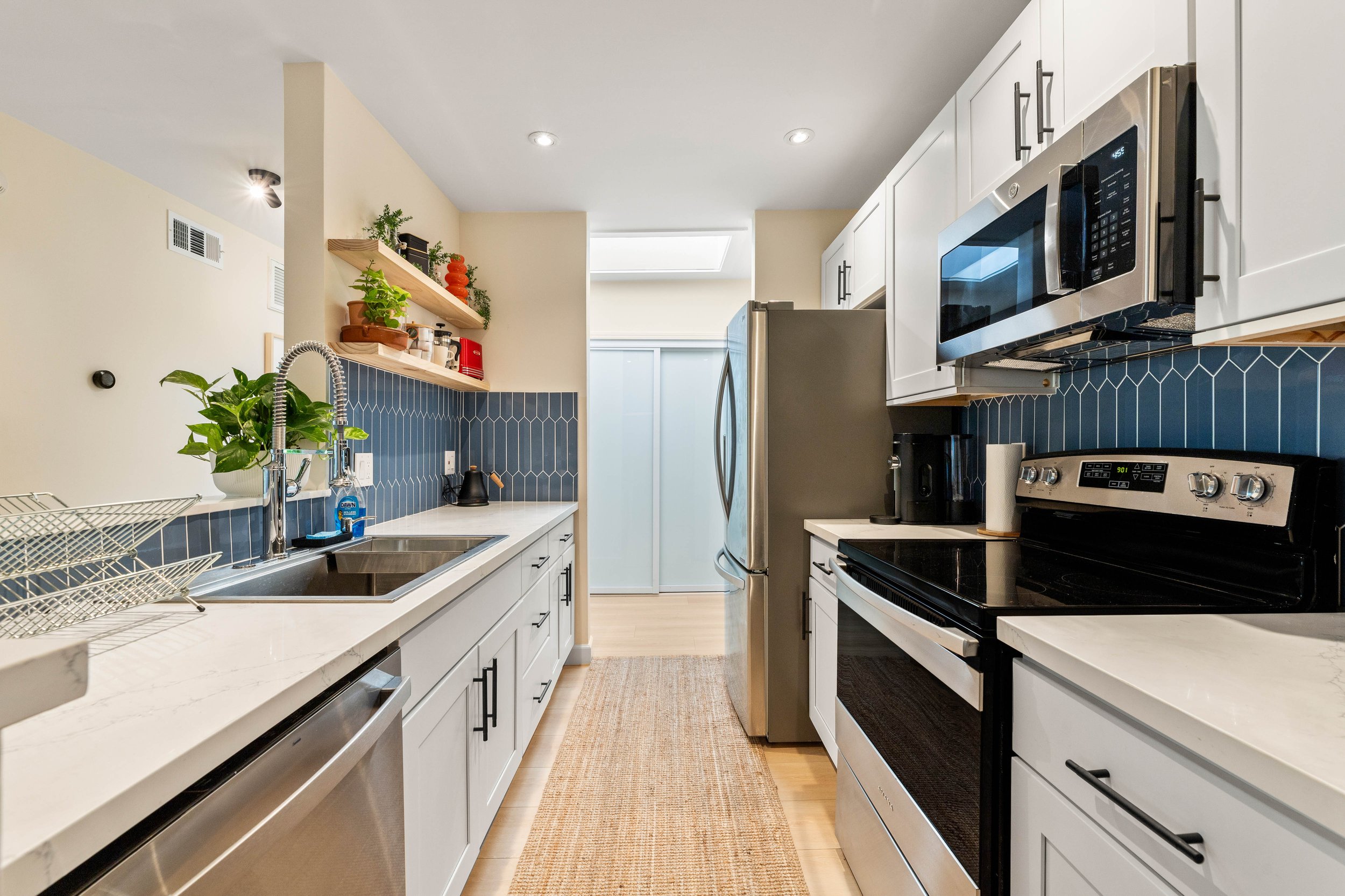 Modern kitchen with white cabinets, black hardware, stainless steel appliances, blue tile backsplash, and a beige rug on light wood flooring.