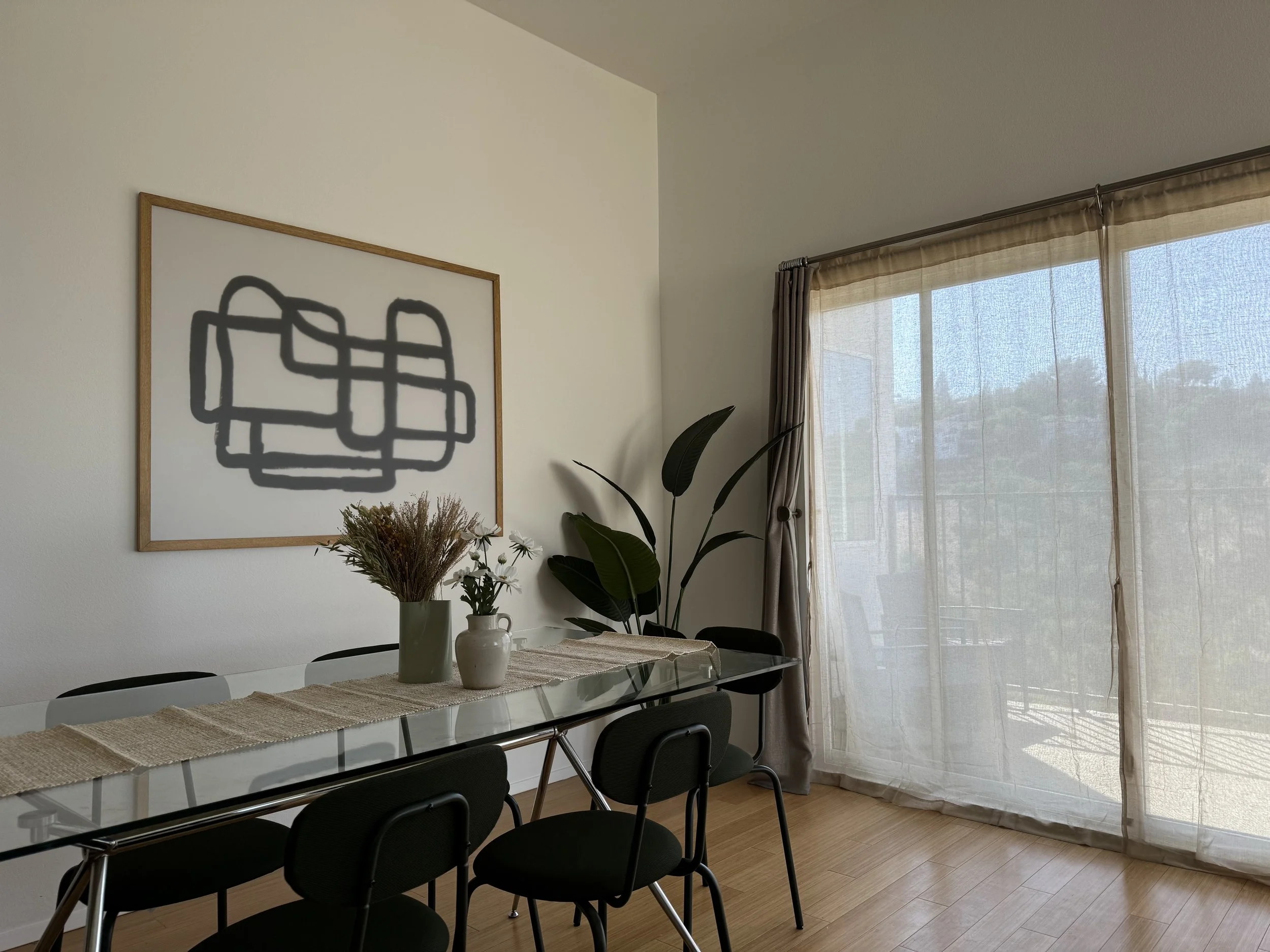A dining room with a glass table, black chairs, and a wall art piece. There's a large window with sheer curtains and some green plants on the table and in the corner.