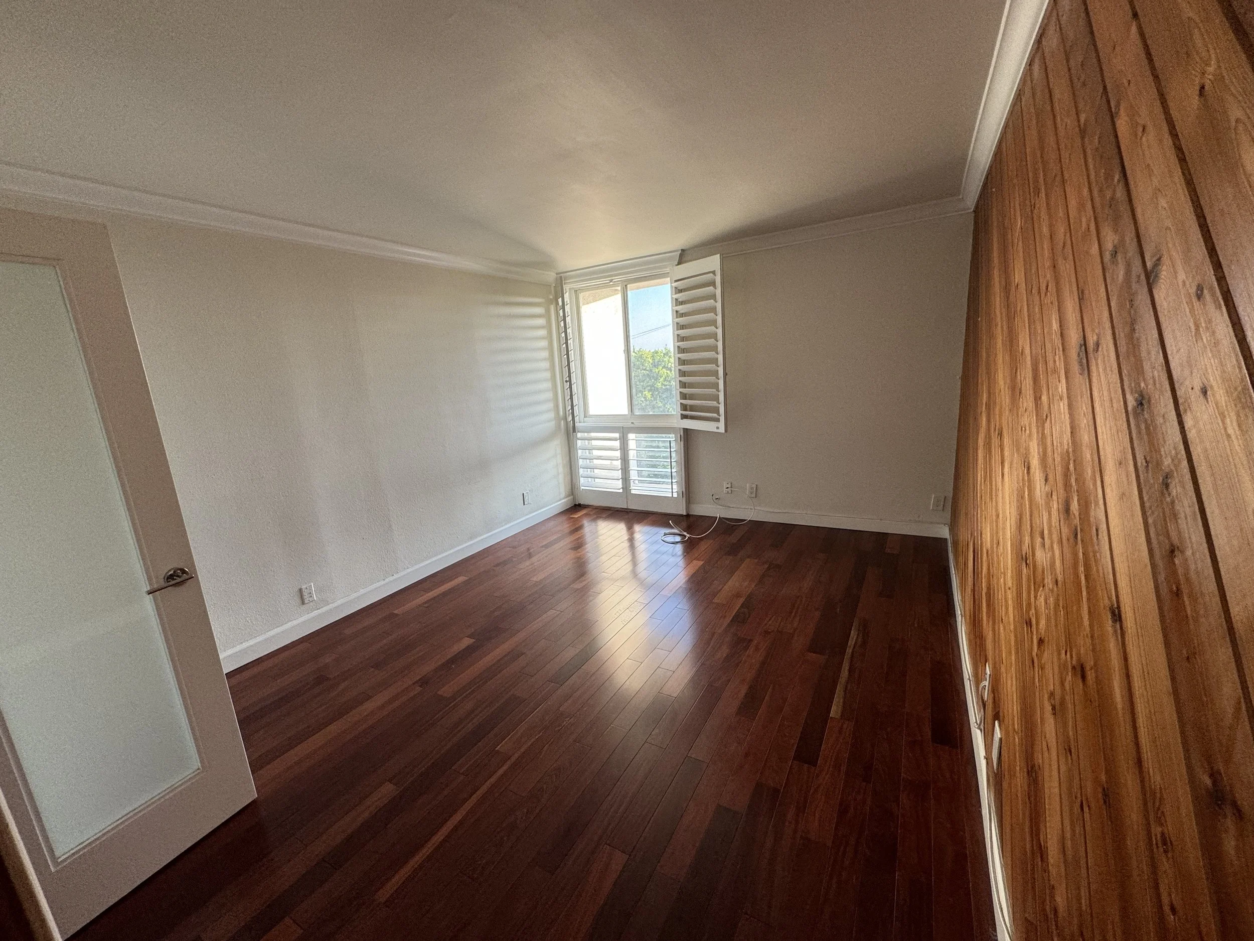 Empty room with hardwood floors, white walls, window with shutters, and some electrical cables on the floor.