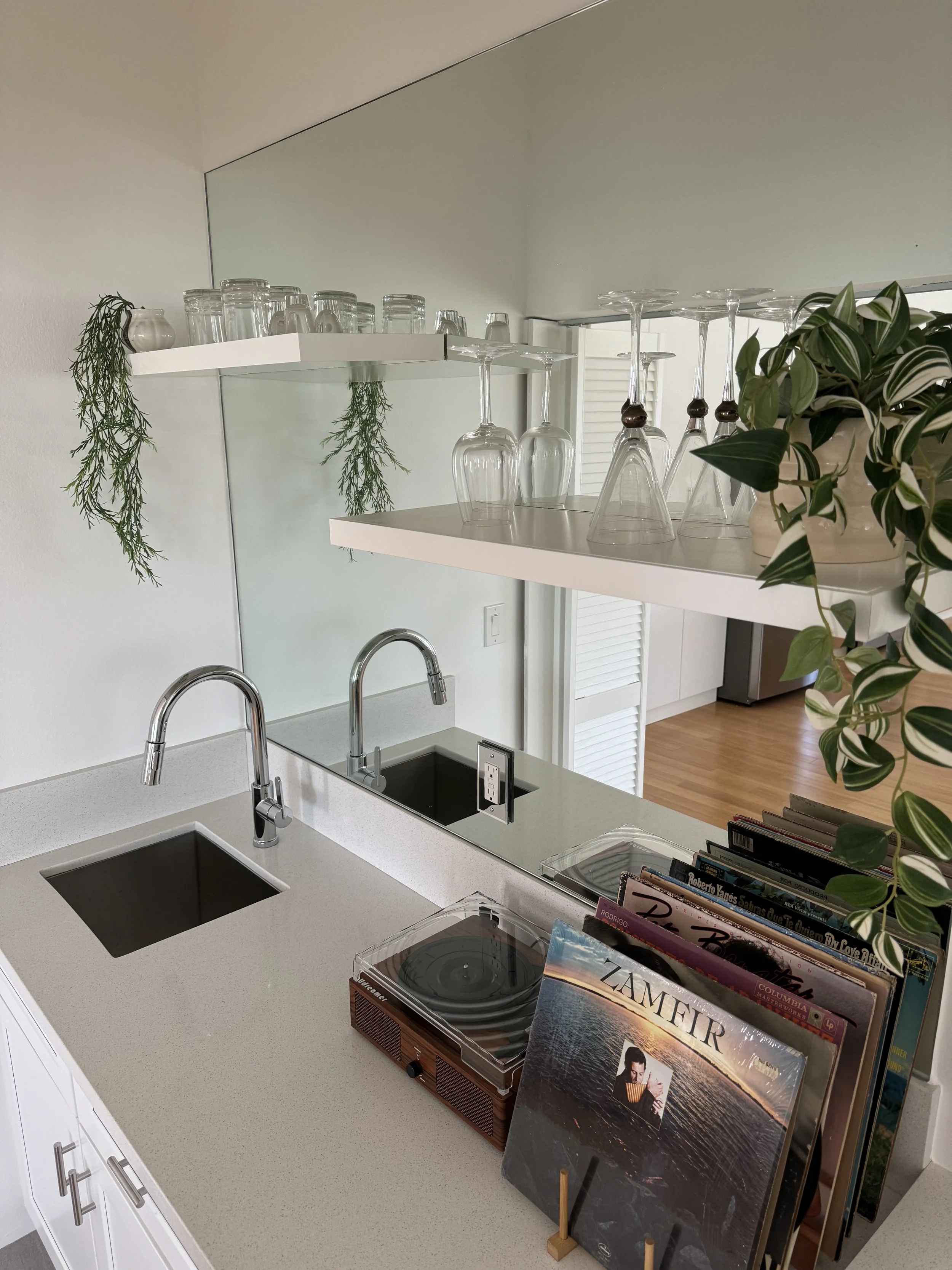 Kitchen counter with a record player, magazines, and a book, with a sink and a faucet in the foreground, and shelves with glasses, plants, and books in the background.