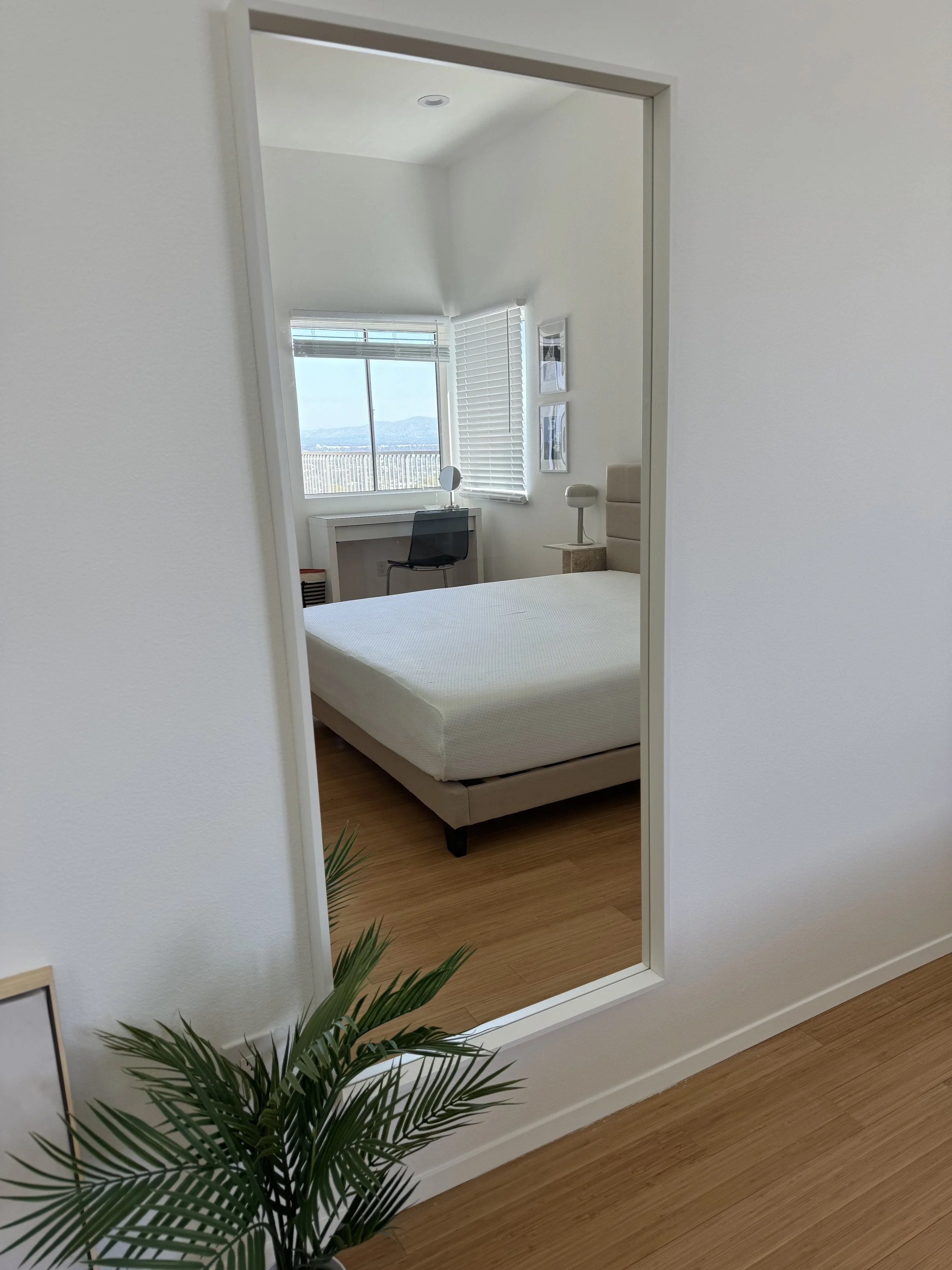 A mirror reflecting a minimalist bedroom with a bed, window, and desk. The room has white walls, wooden flooring, and window blinds. There is a potted plant in the foreground.
