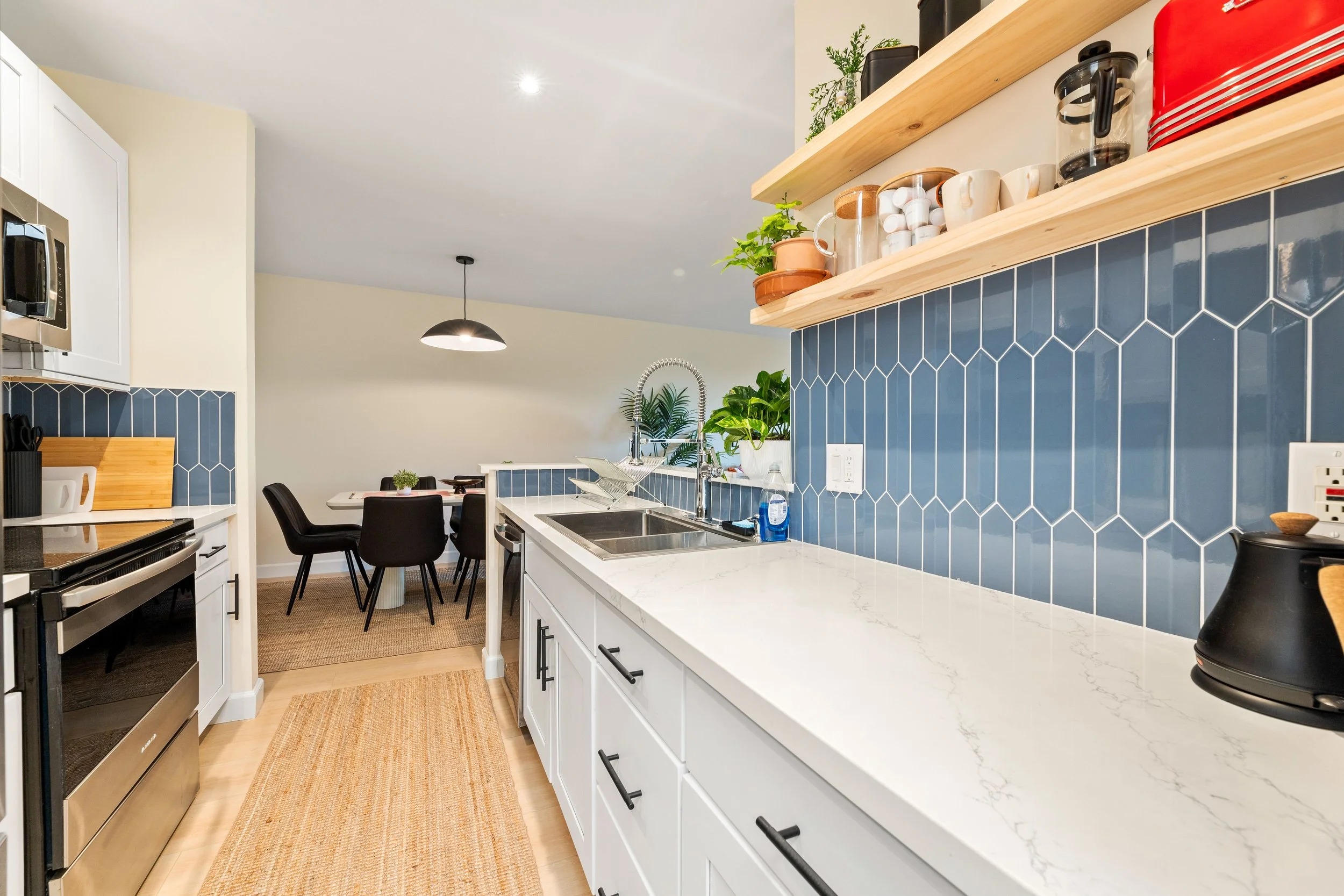 A modern kitchen with white cabinets, a white marble countertop, dark blue tiled backsplash, and open wooden shelves displaying plants and kitchenware. The kitchen has a sink with a stainless steel faucet, and adjacent is a dining area with a round table and black chairs. Recessed lighting and a black pendant light illuminate the space.